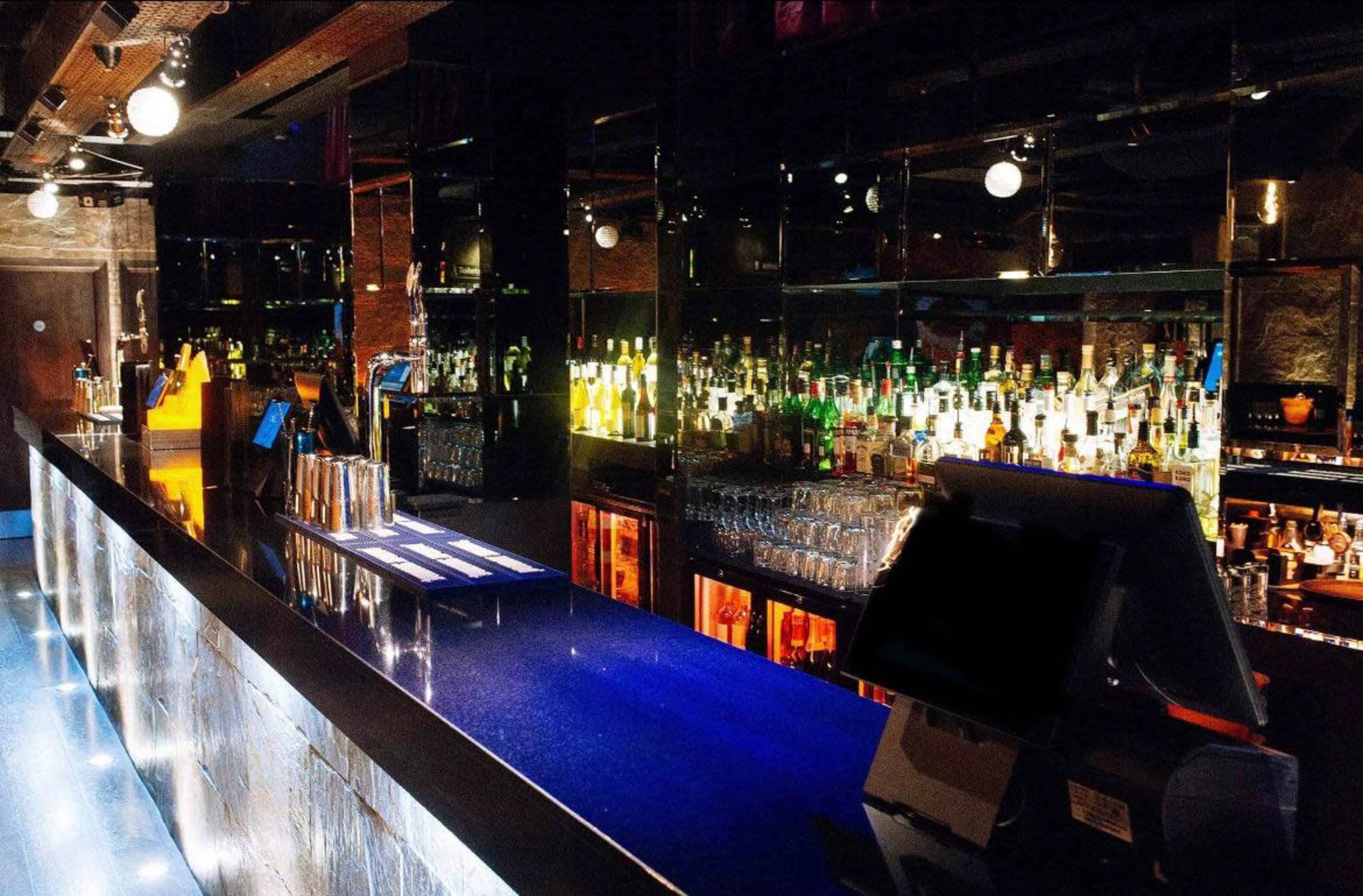The image shows a modern bar with a long, glossy counter, illuminated shelves filled with various bottles, and a sleek, contemporary design.