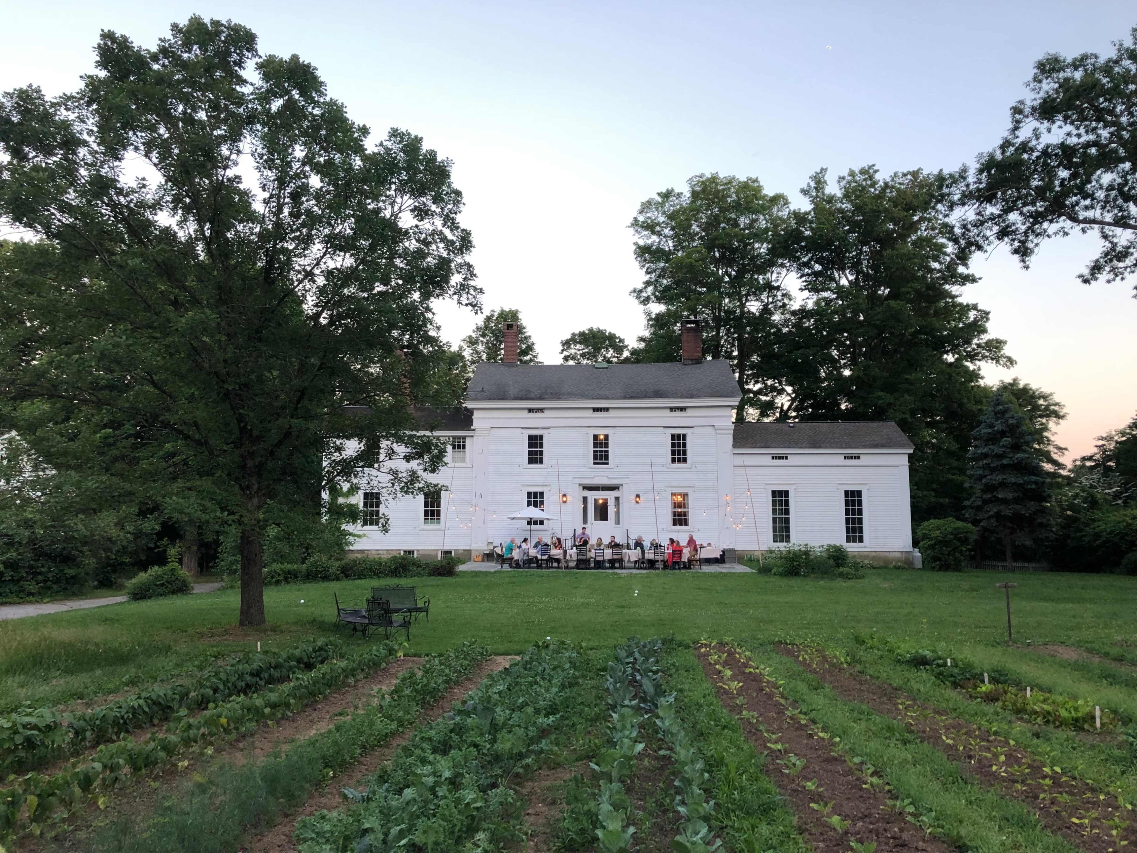 A large white house with a green lawn and rows of vegetables in the foreground hosts a gathering of people on its porch during twilight.