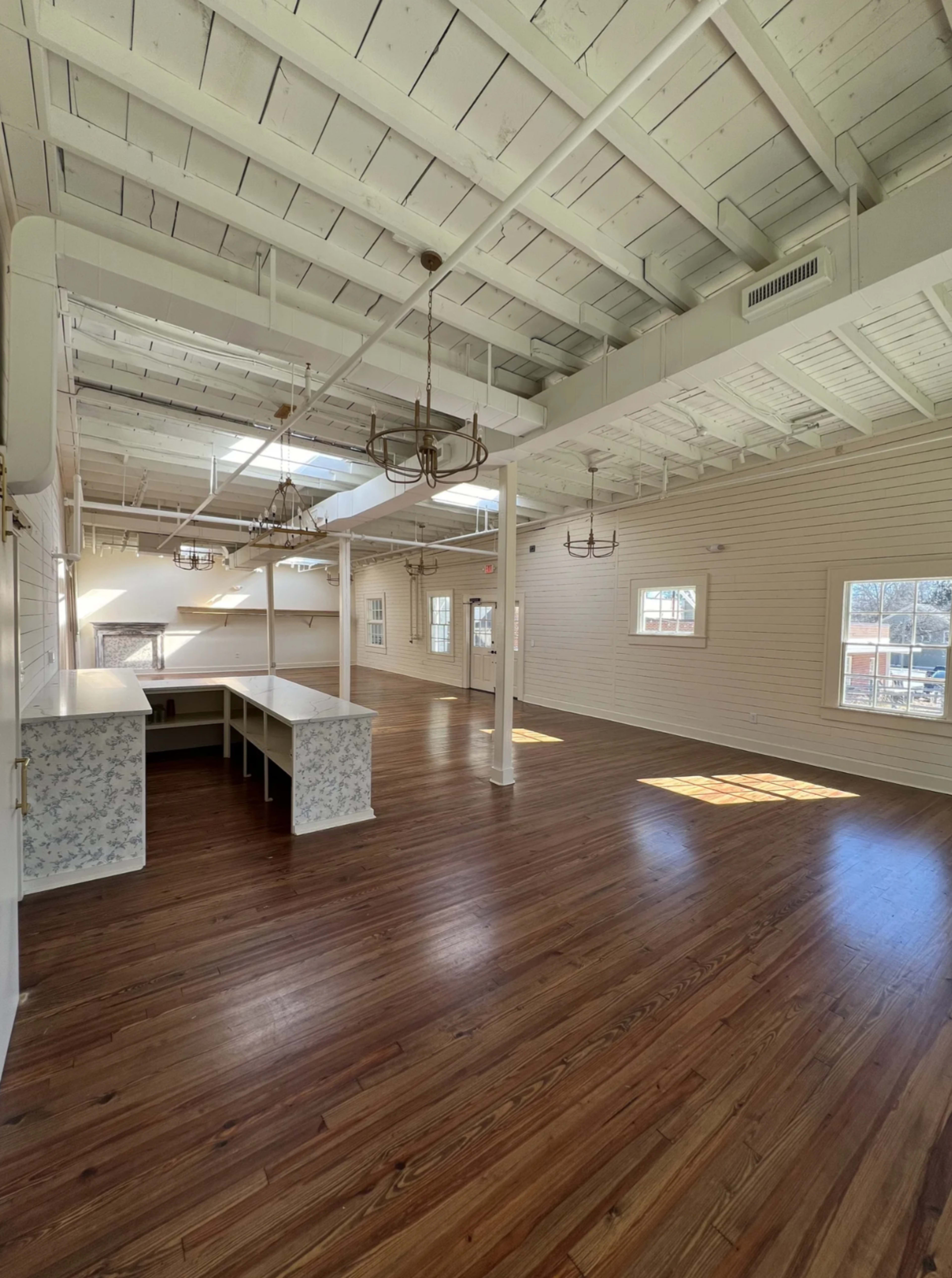 The image depicts a spacious, empty room with high ceilings, hardwood floors, and large windows allowing natural light to enter.