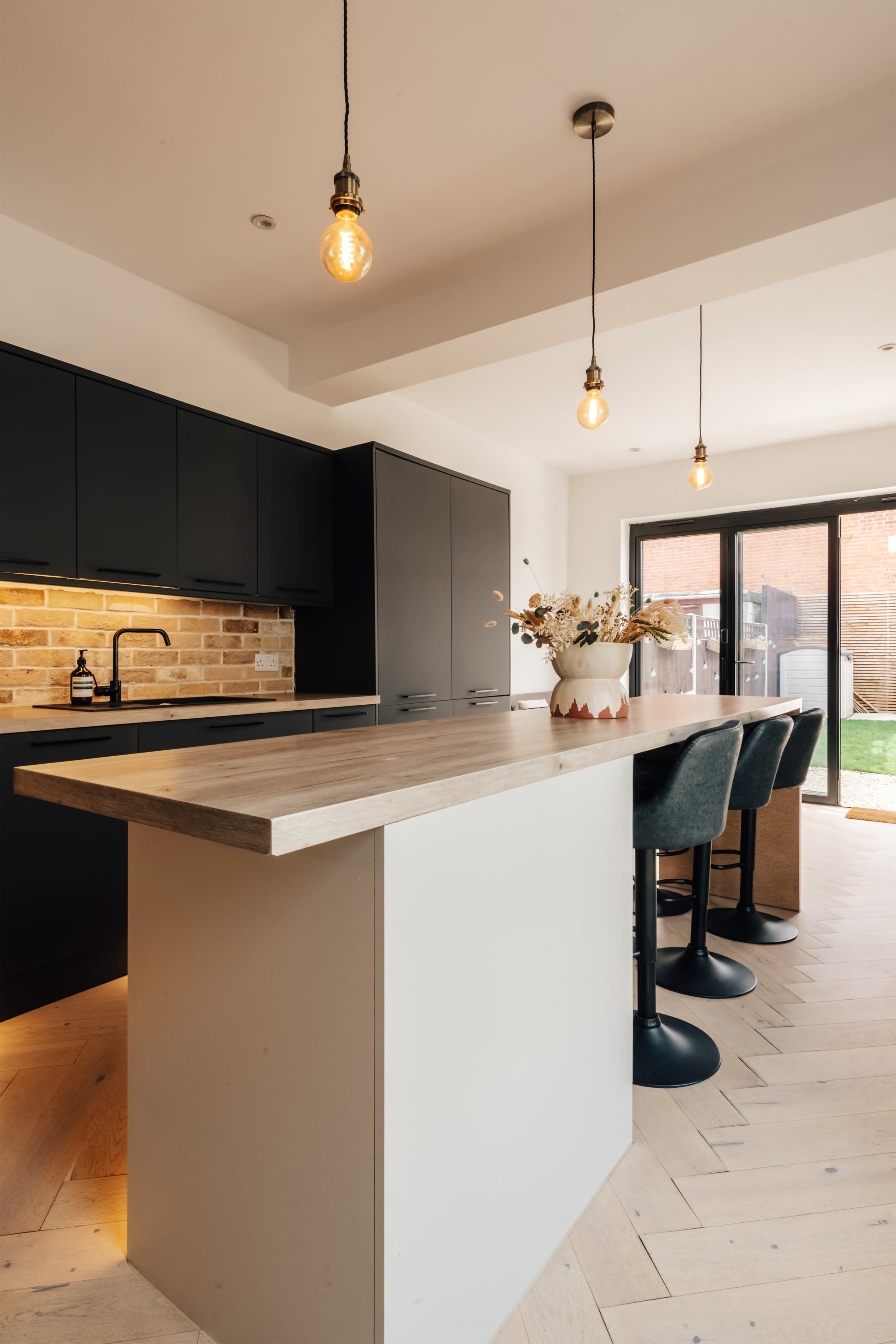 The image shows a modern kitchen featuring dark cabinetry, a light wooden countertop, and three bar stools under pendant lighting.