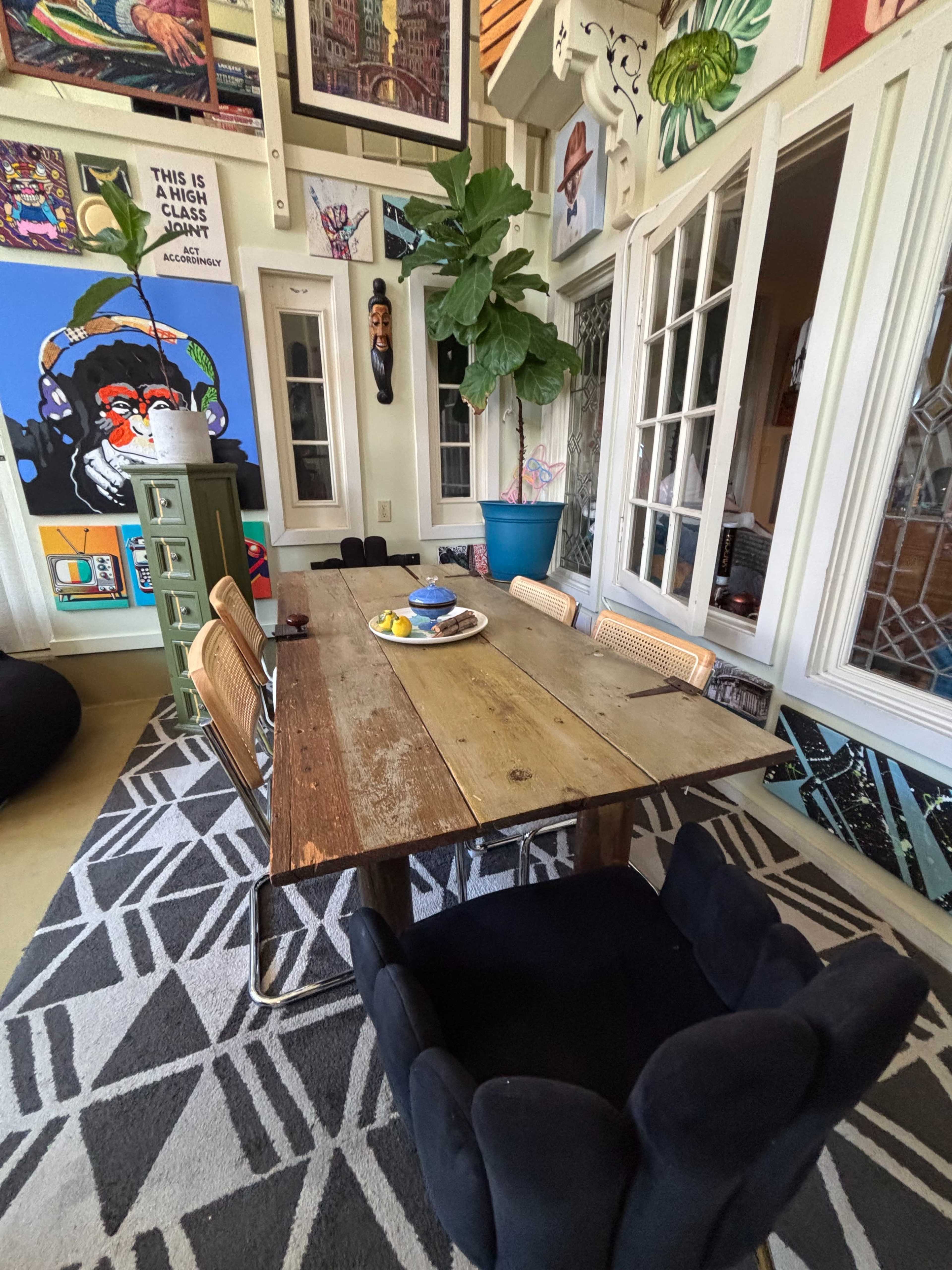 The image shows a dining area with a wooden table surrounded by chairs, colorful artwork on the walls, and a large plant in a blue pot.
