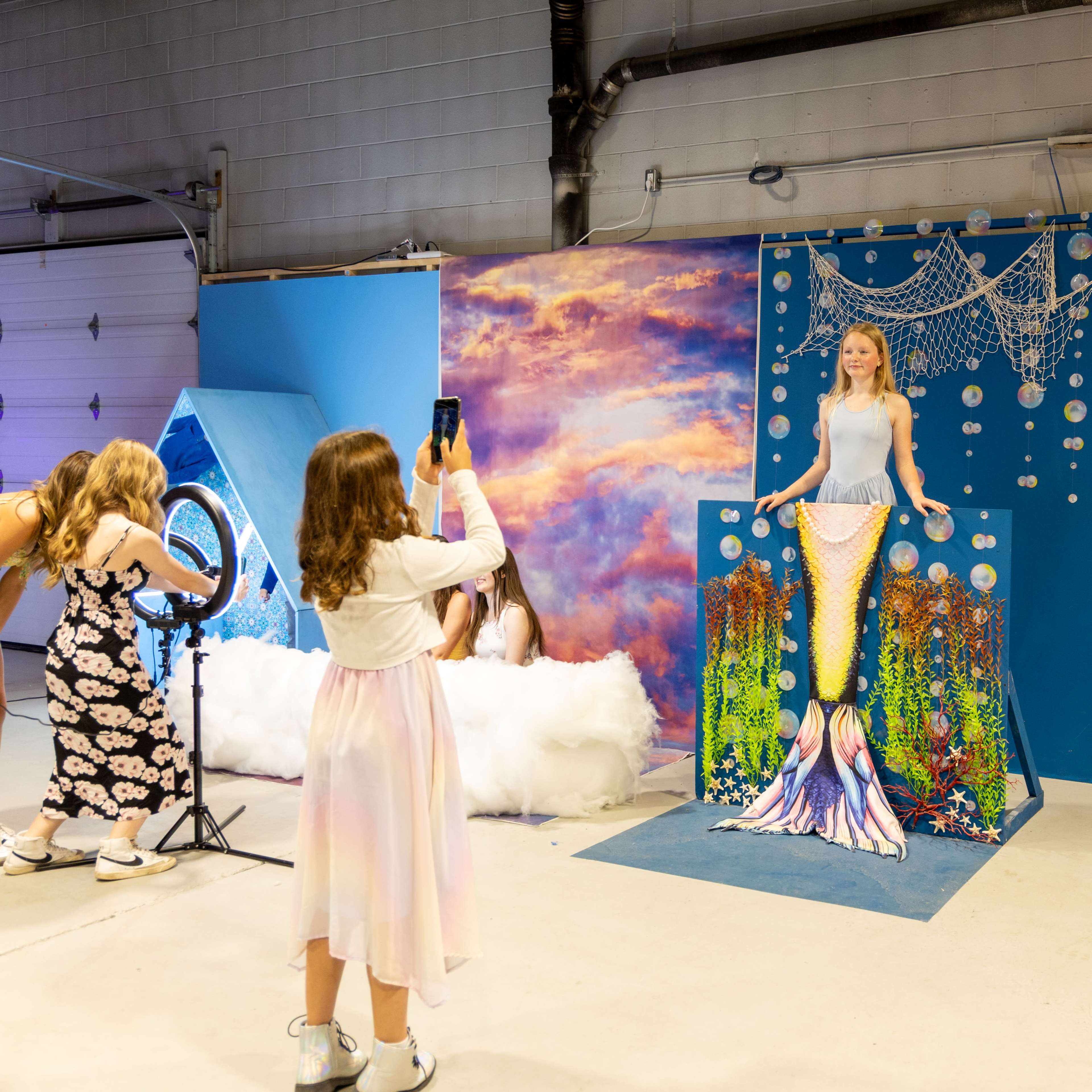A group of girls is taking photos in front of a mermaid-themed backdrop in a brightly lit indoor space.