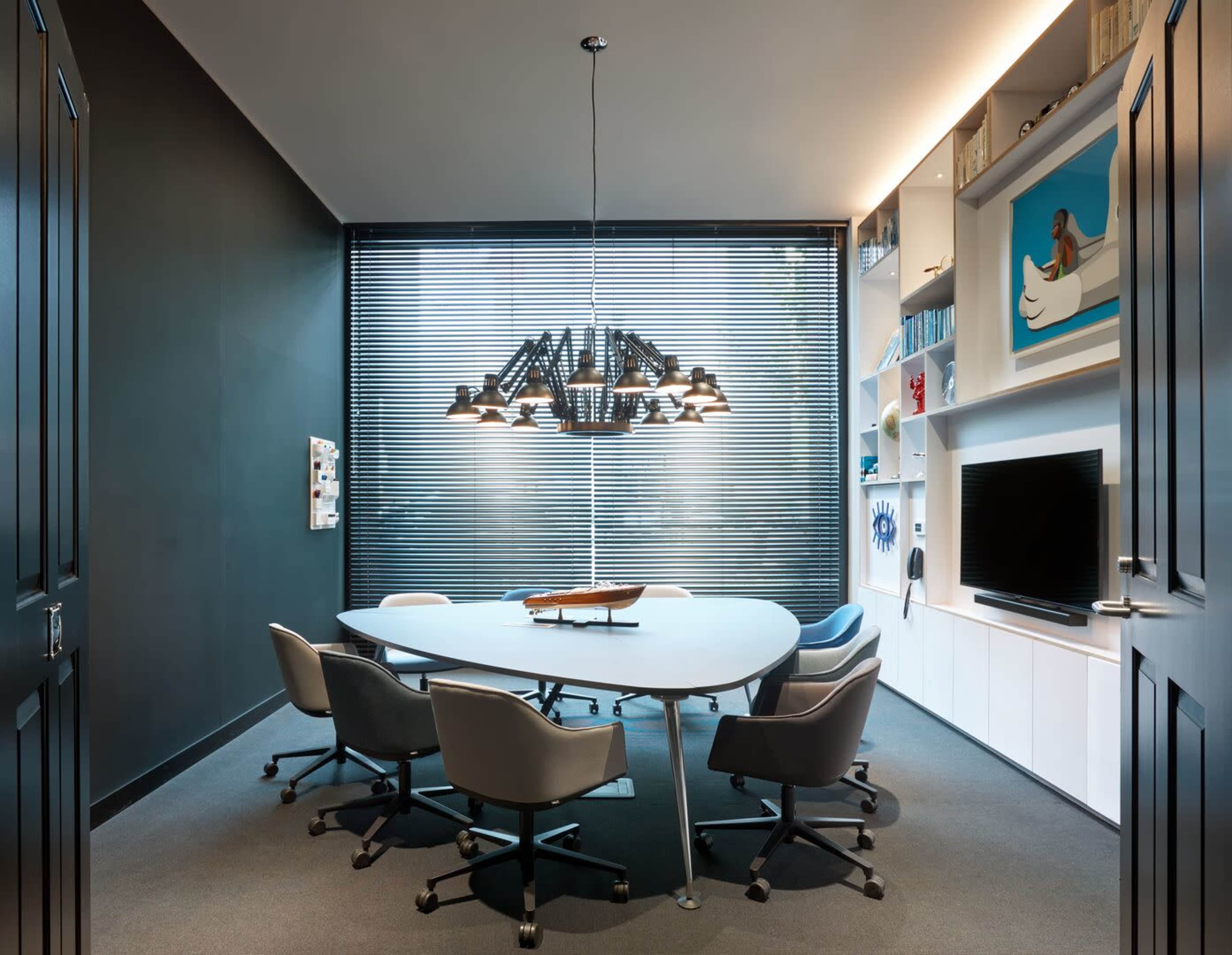 A modern conference room features a round table surrounded by six chairs, with a large pendant light overhead and shelves displaying decor, set against dark walls and window blinds.