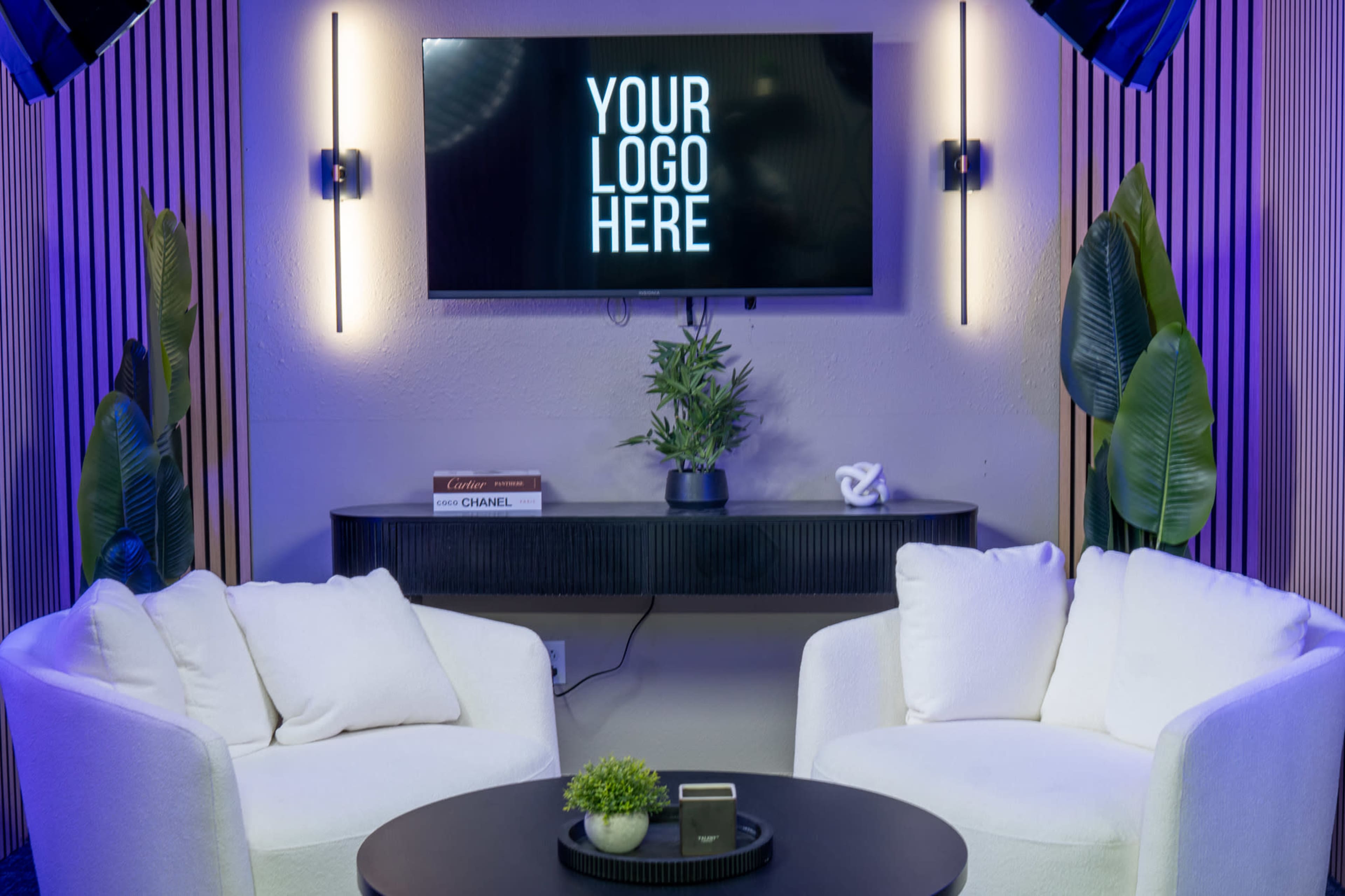 The image shows a modern display area with two white armchairs facing a circular black table, a small plant on the table, and a television mounted on the wall featuring placeholder text for a logo.