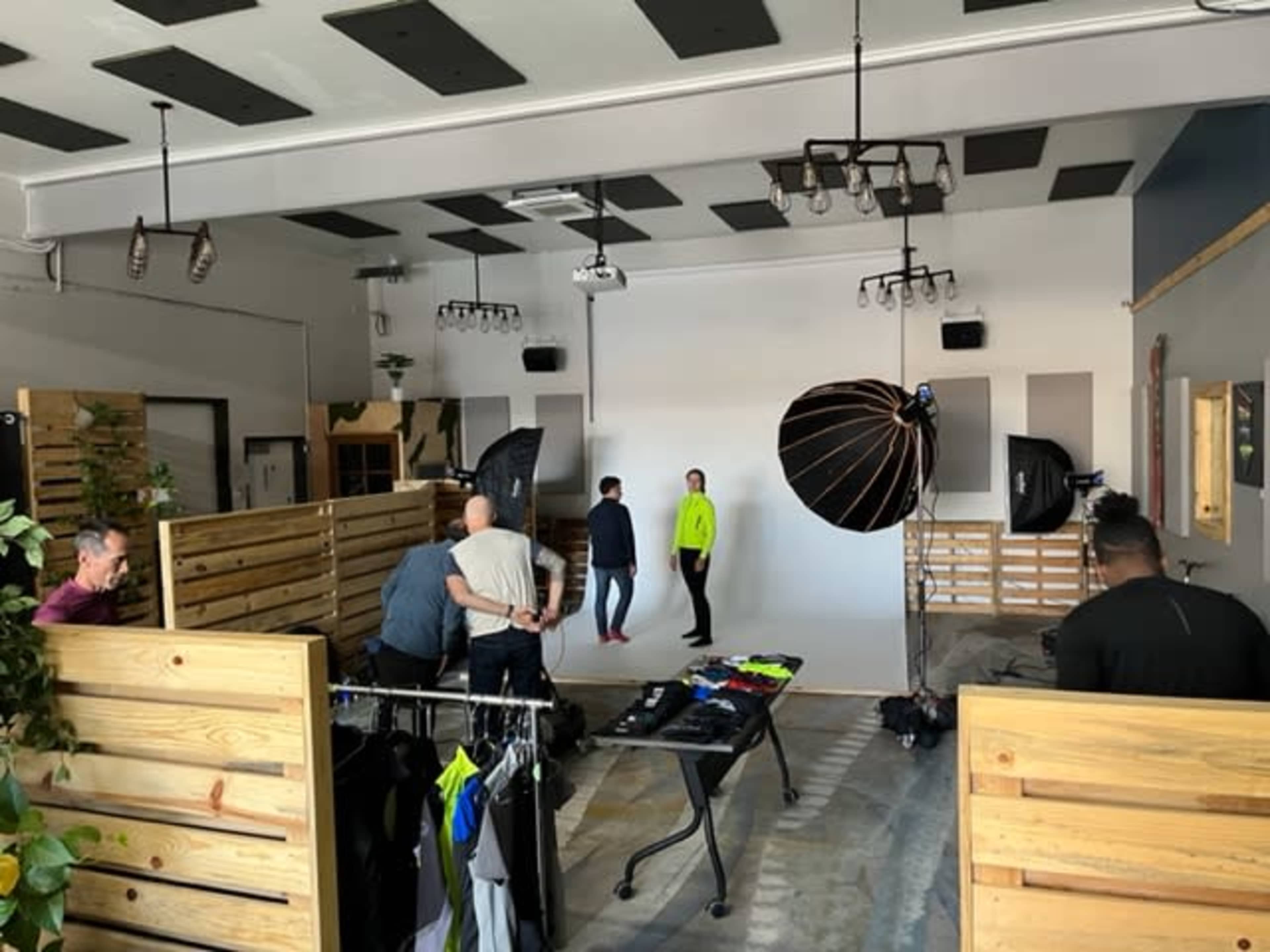 A photography studio is set up with two models posing against a white backdrop, surrounded by lighting equipment and clothing racks.