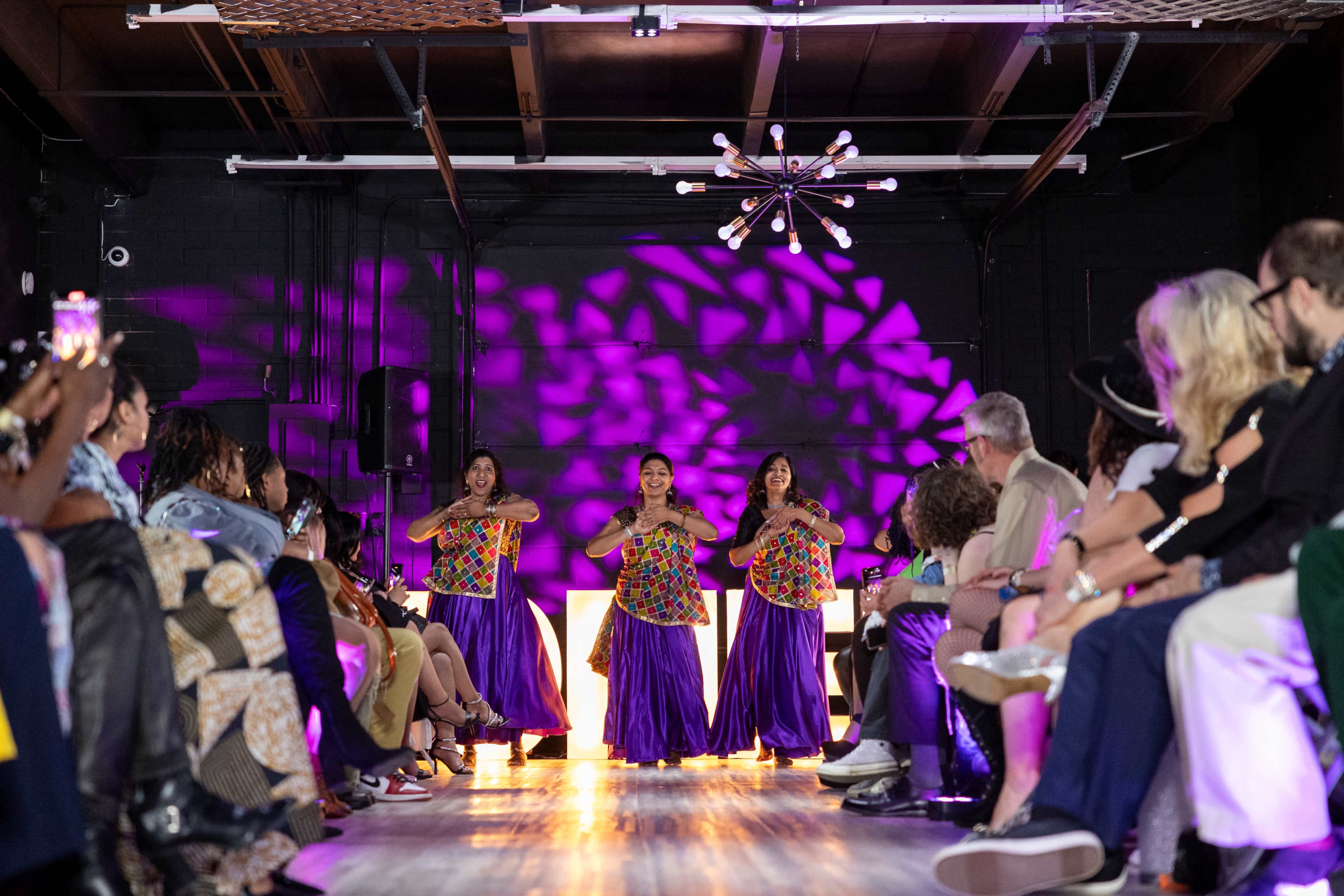 Three dancers perform in colorful attire on a stage illuminated by purple lights, while an audience watches from the sides.