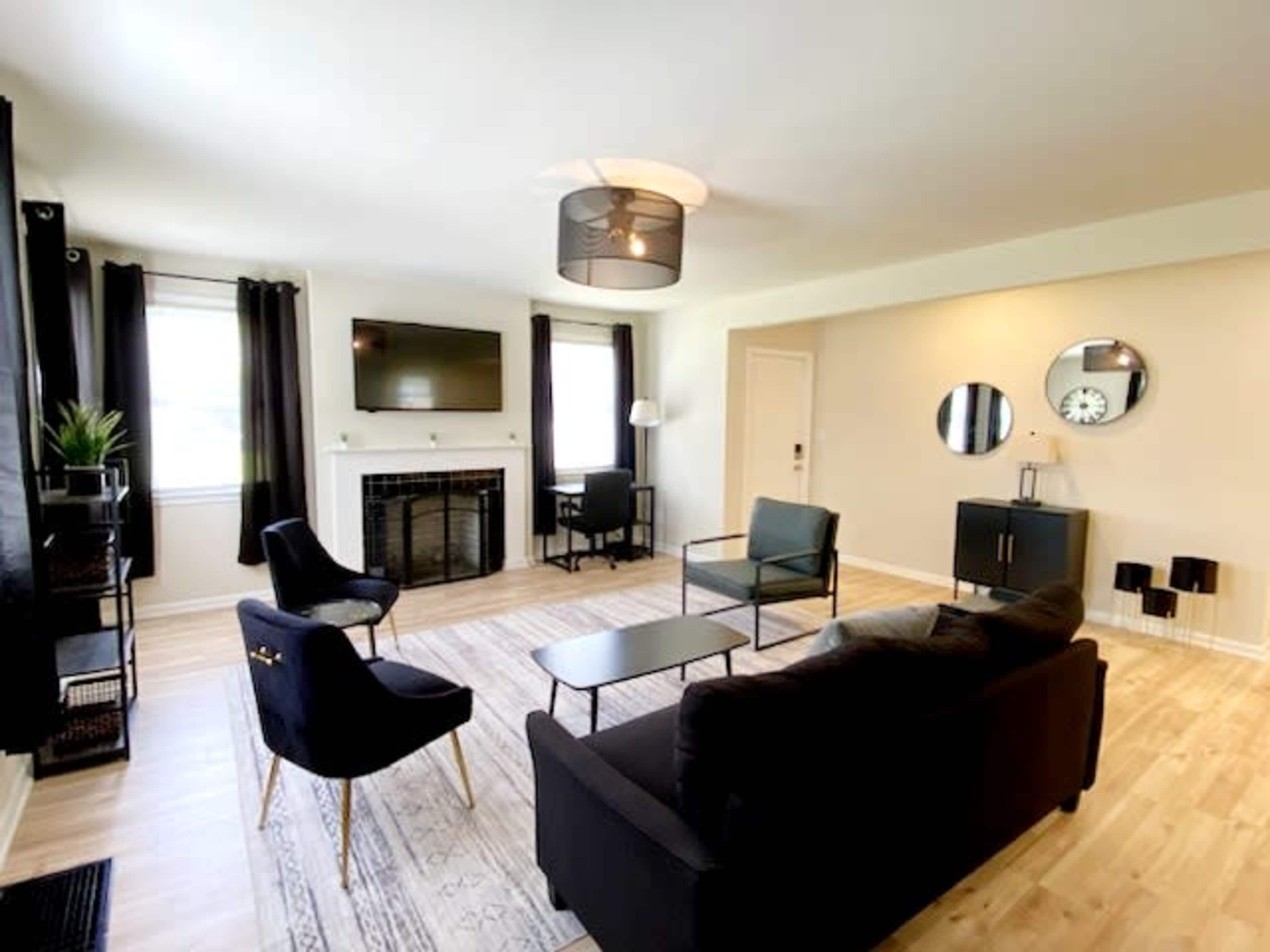 The living room features a central black sofa, two chairs, a coffee table, a fireplace, and a flat-screen TV mounted on the wall, with large windows dressed in dark curtains.