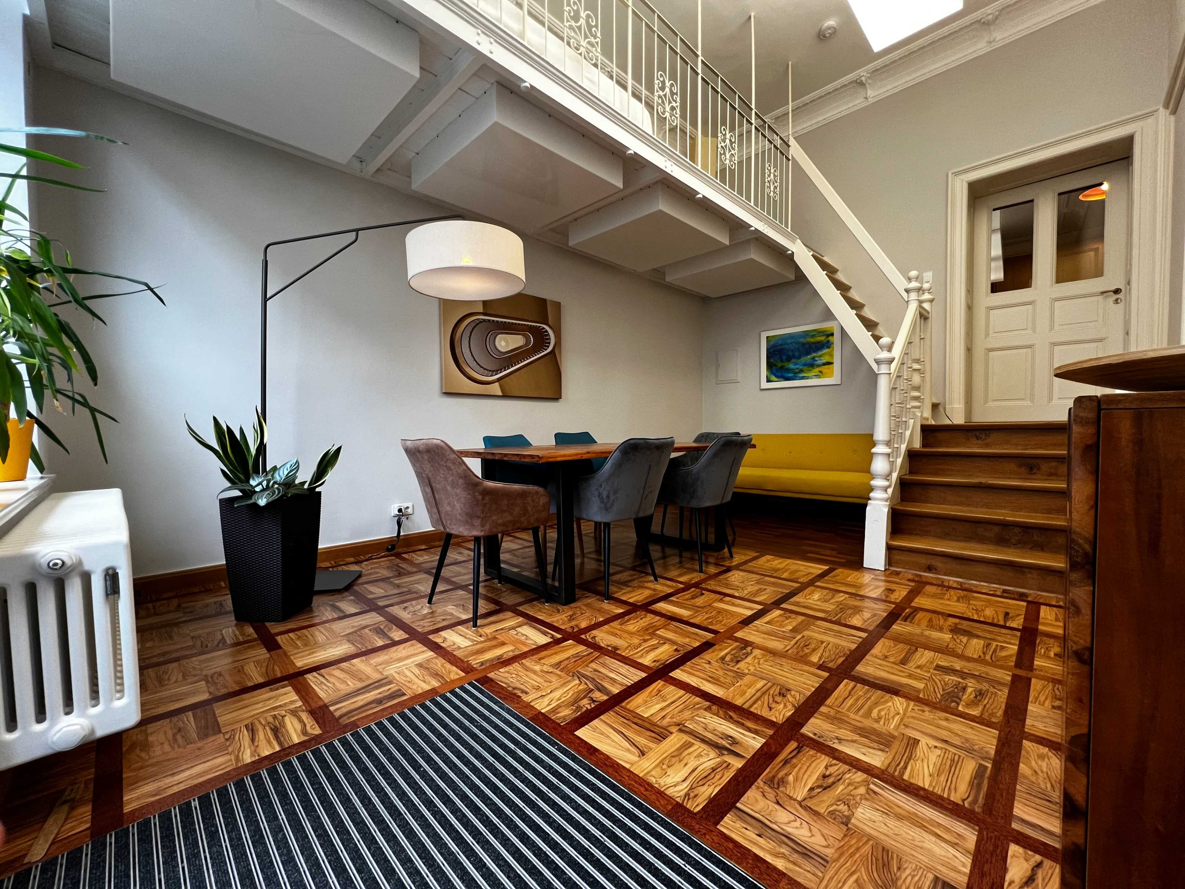 The image shows a spacious interior with a dining table surrounded by chairs, a staircase, and large windows, enhanced by wooden flooring and modern decor.