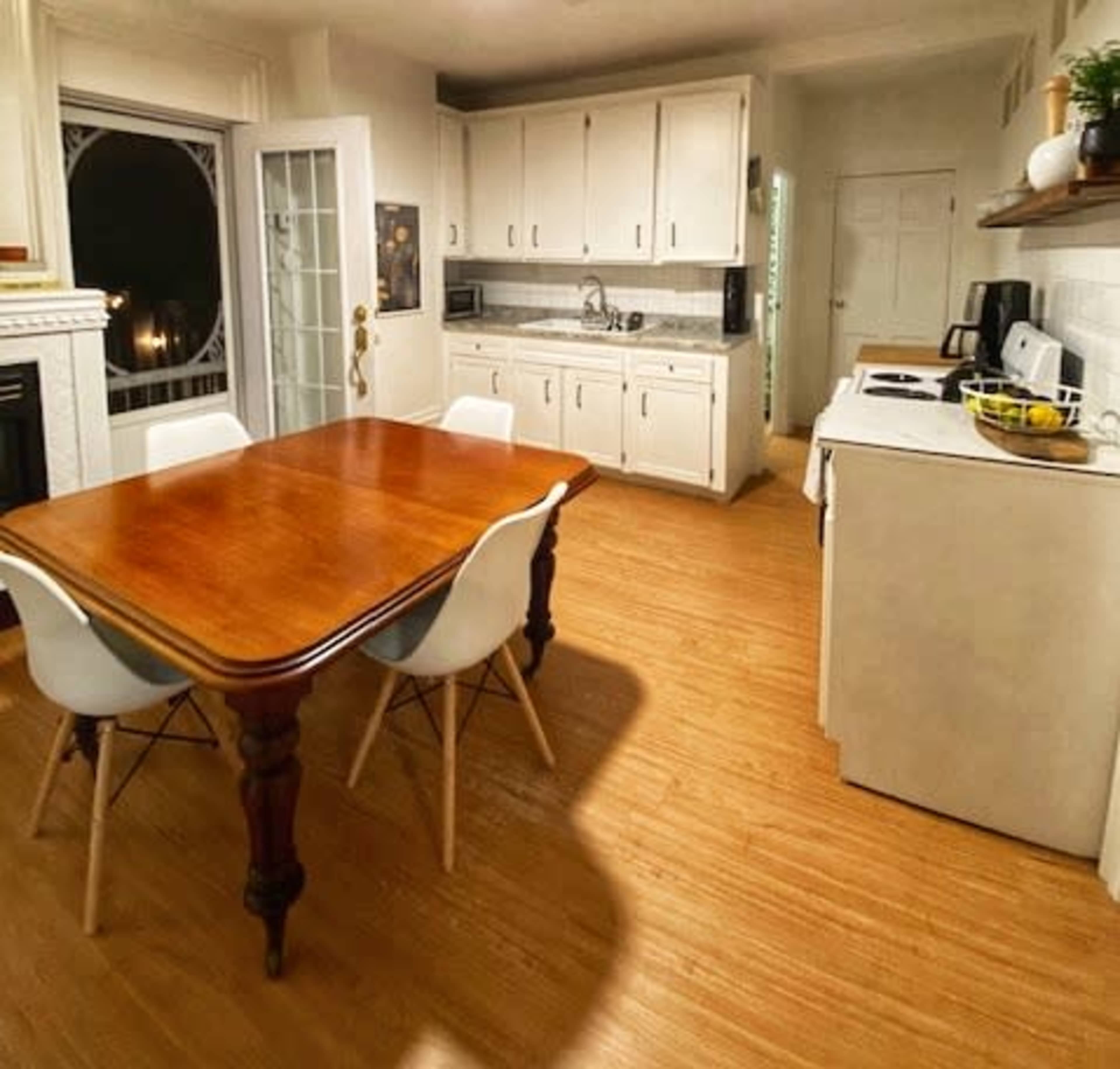 A kitchen with a wooden dining table and white chairs, featuring a stove, refrigerator, and cabinets in a well-lit space.