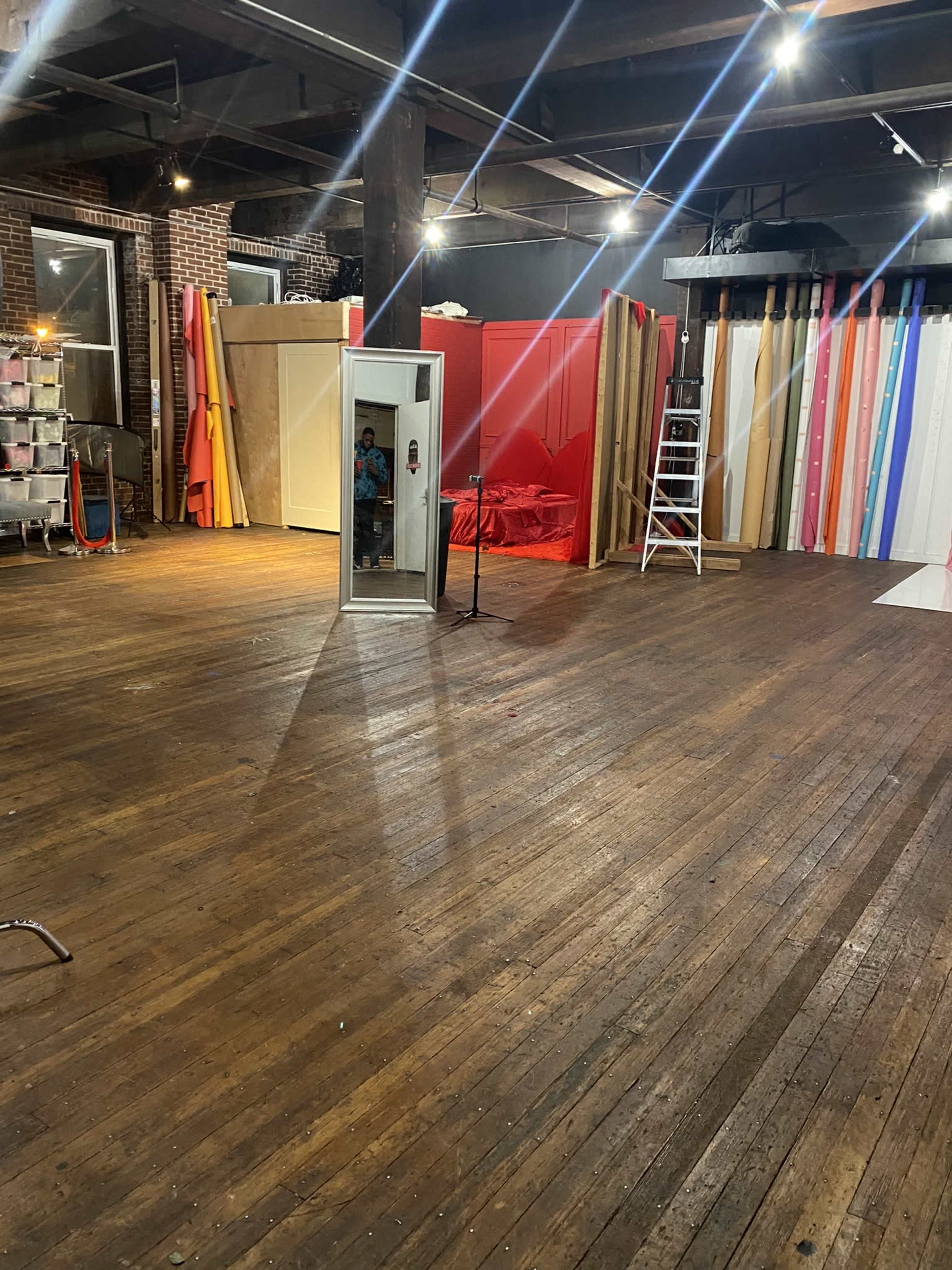 The image shows a spacious studio with wooden floors, a large mirror, a microphone stand, and colorful backdrops arranged along one wall.