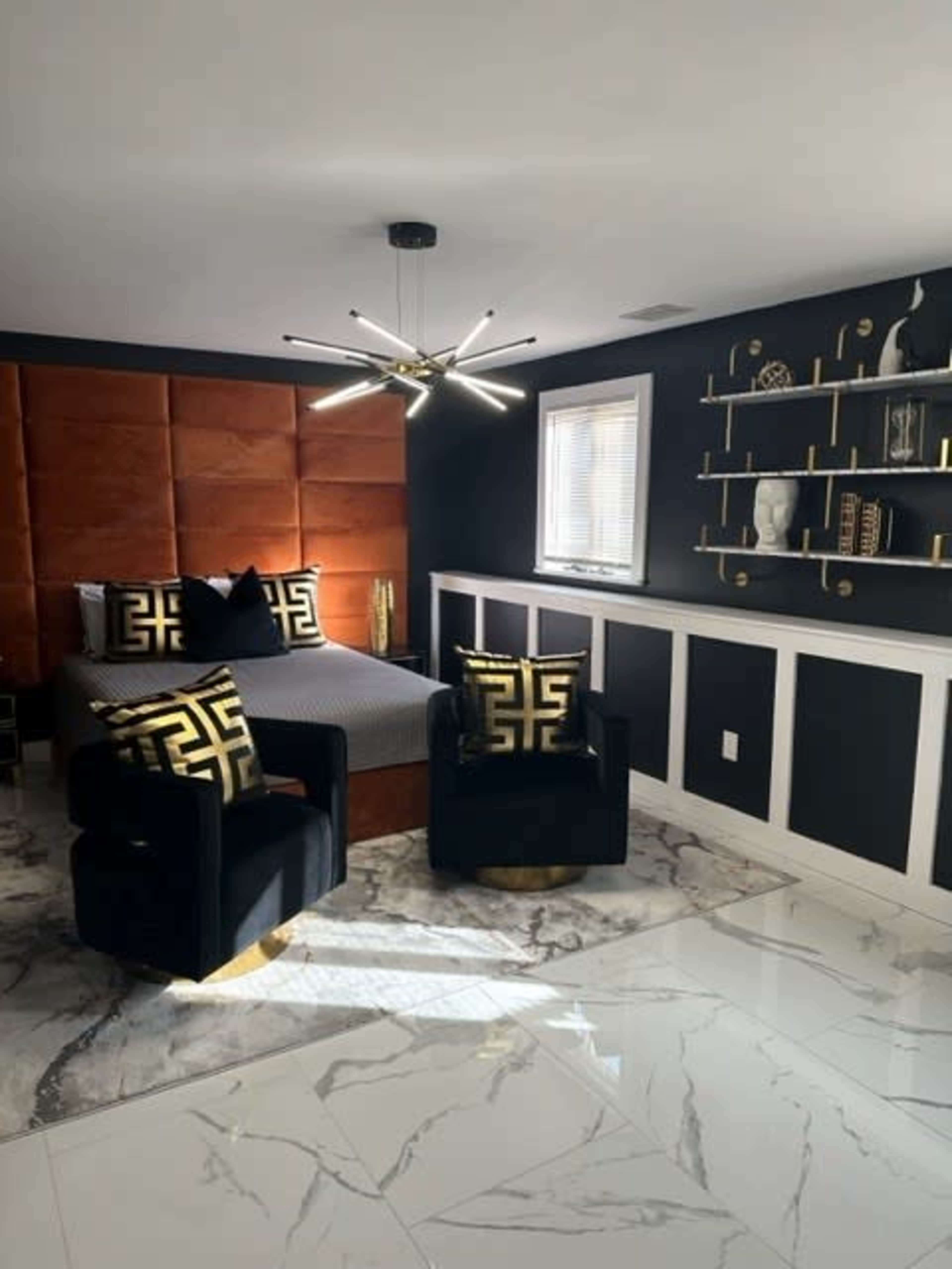 A modern bedroom featuring a bed with a tufted brown headboard, two black velvet chairs with gold accents, and a sleek bookshelf against a dark wall.