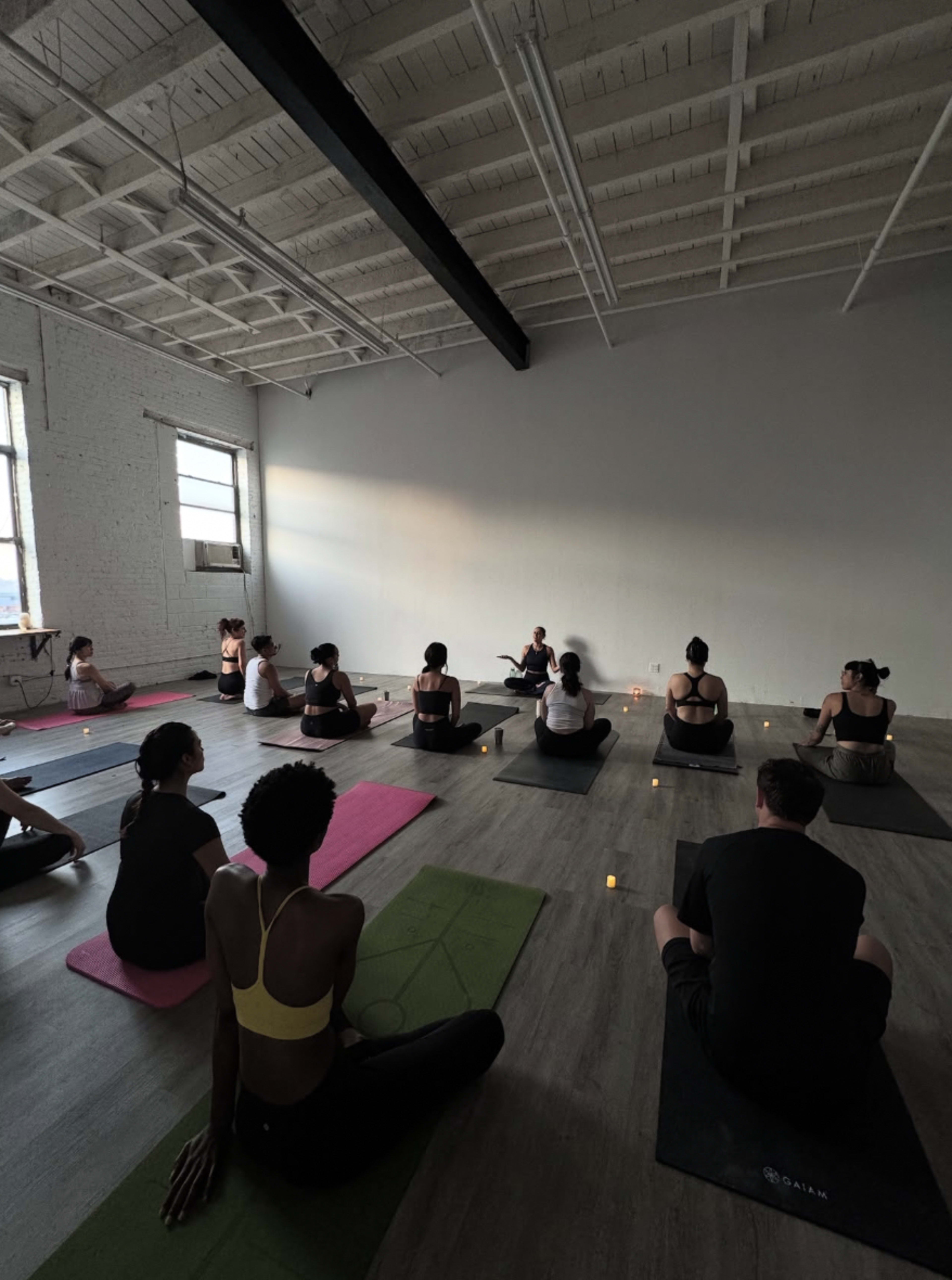 A group of people sits on yoga mats in a dimly lit room, facing an instructor seated at the front, with candles placed around them.