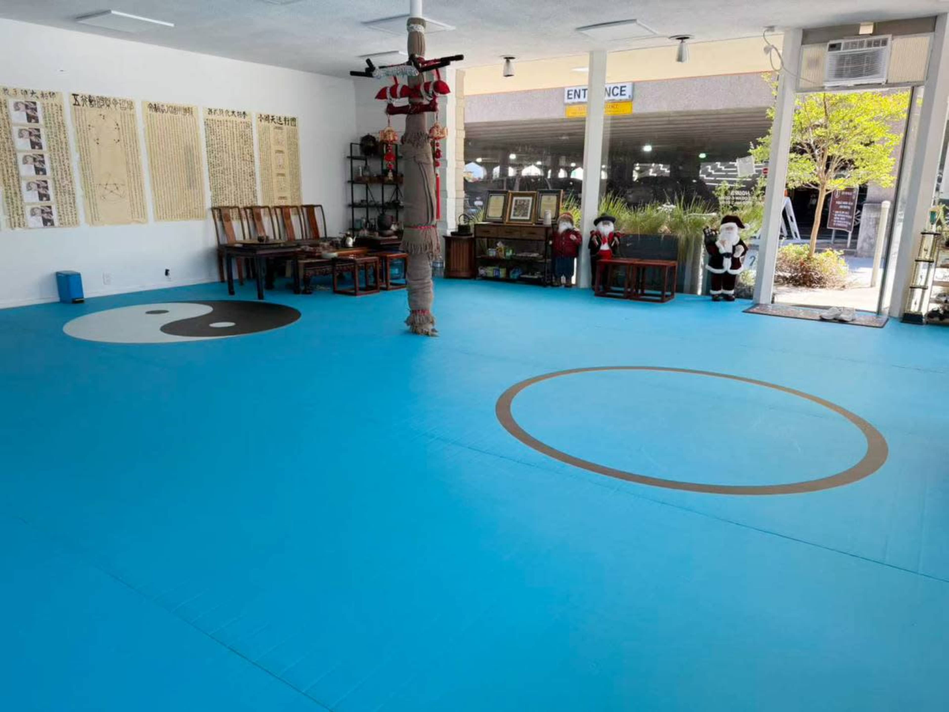 The image shows a brightly lit dojo with a blue mat, featuring a yin-yang symbol and a circular design on the floor, along with wooden furniture and decorations around the edges.