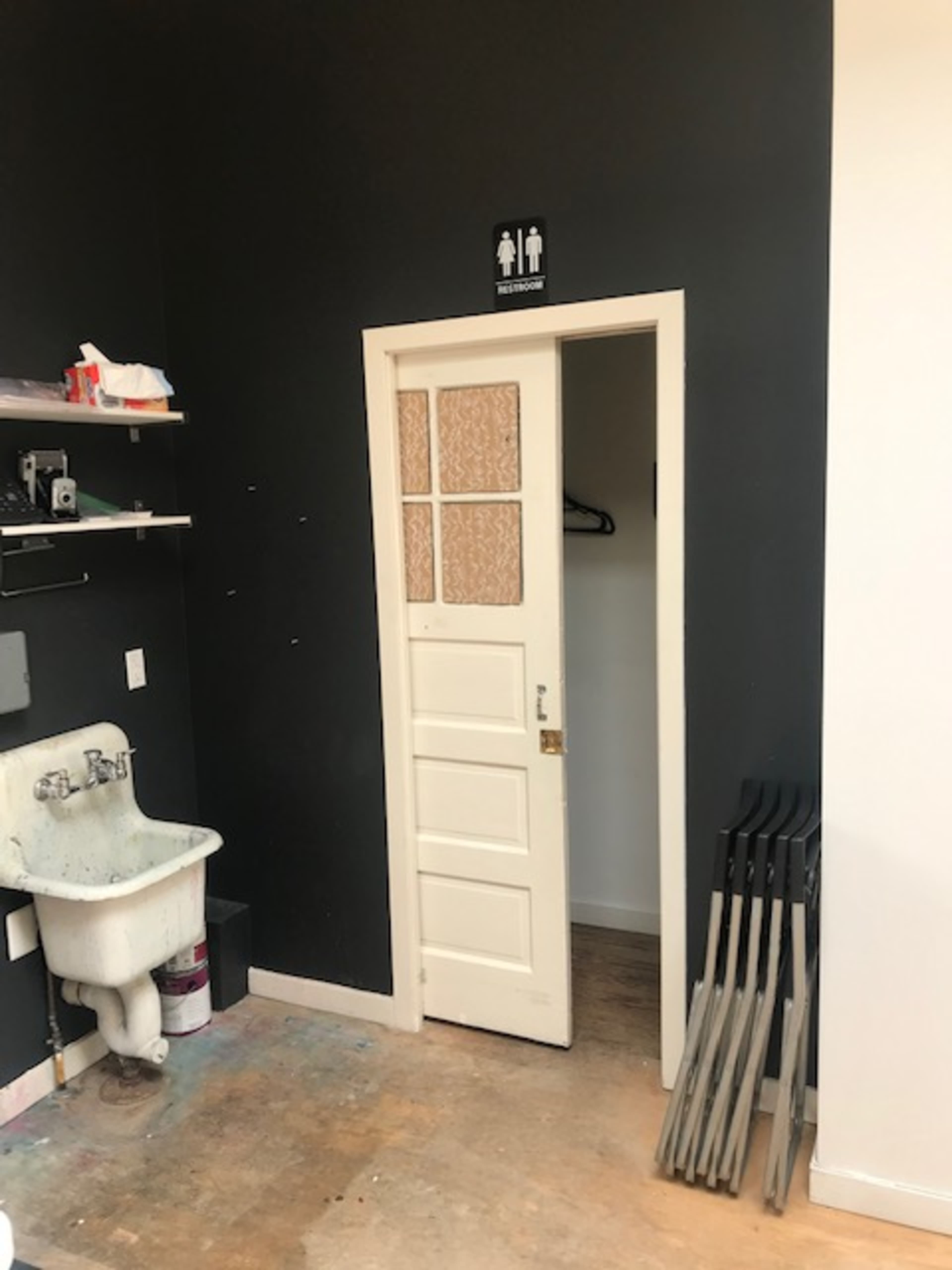 The image shows a white door with a window panel leading to a restroom, surrounded by dark walls, a sink on one side, and several metal implements stacked against the wall.