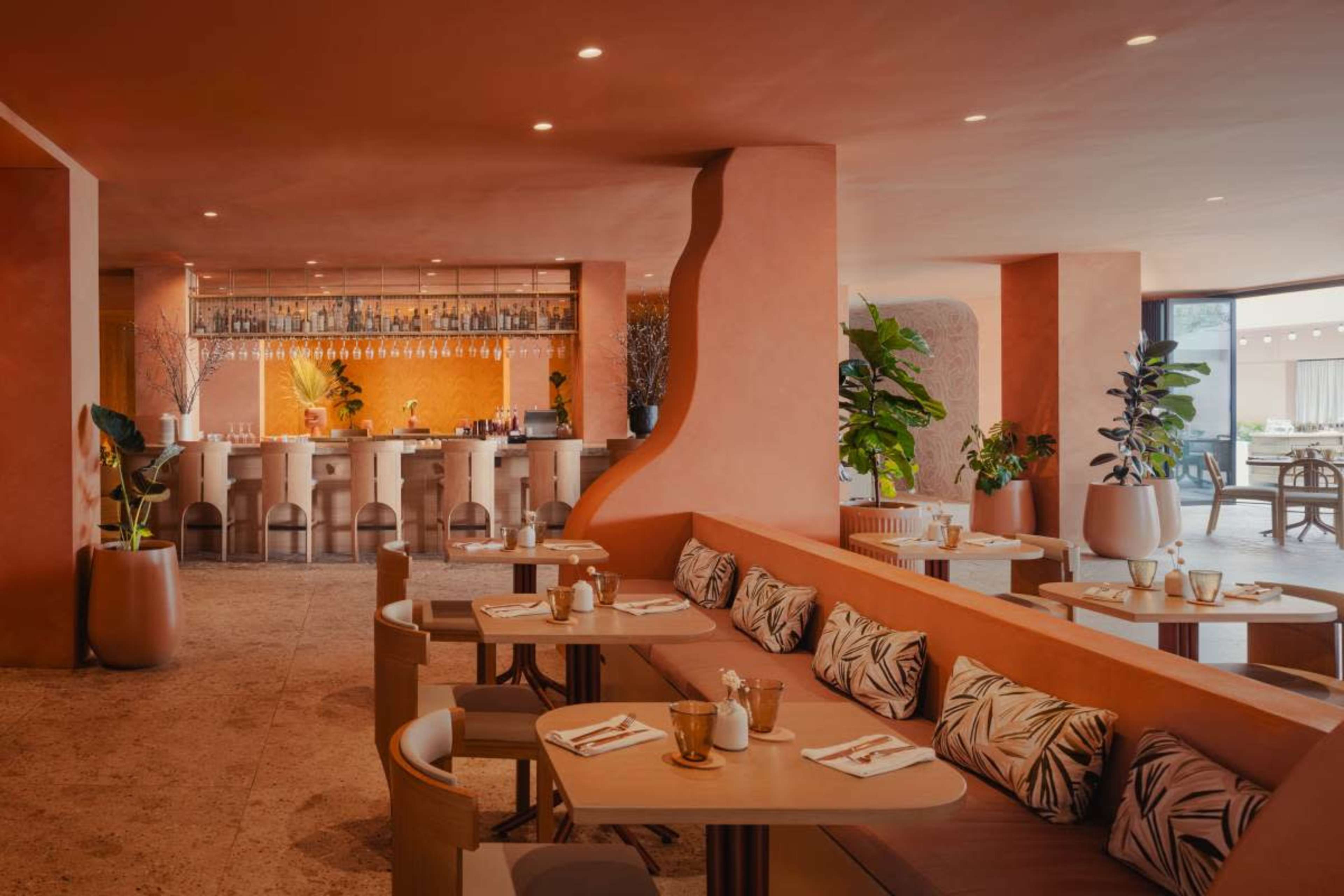 The image shows a modern restaurant interior featuring a bar with a warm-toned design, seating areas, and decorative plants.