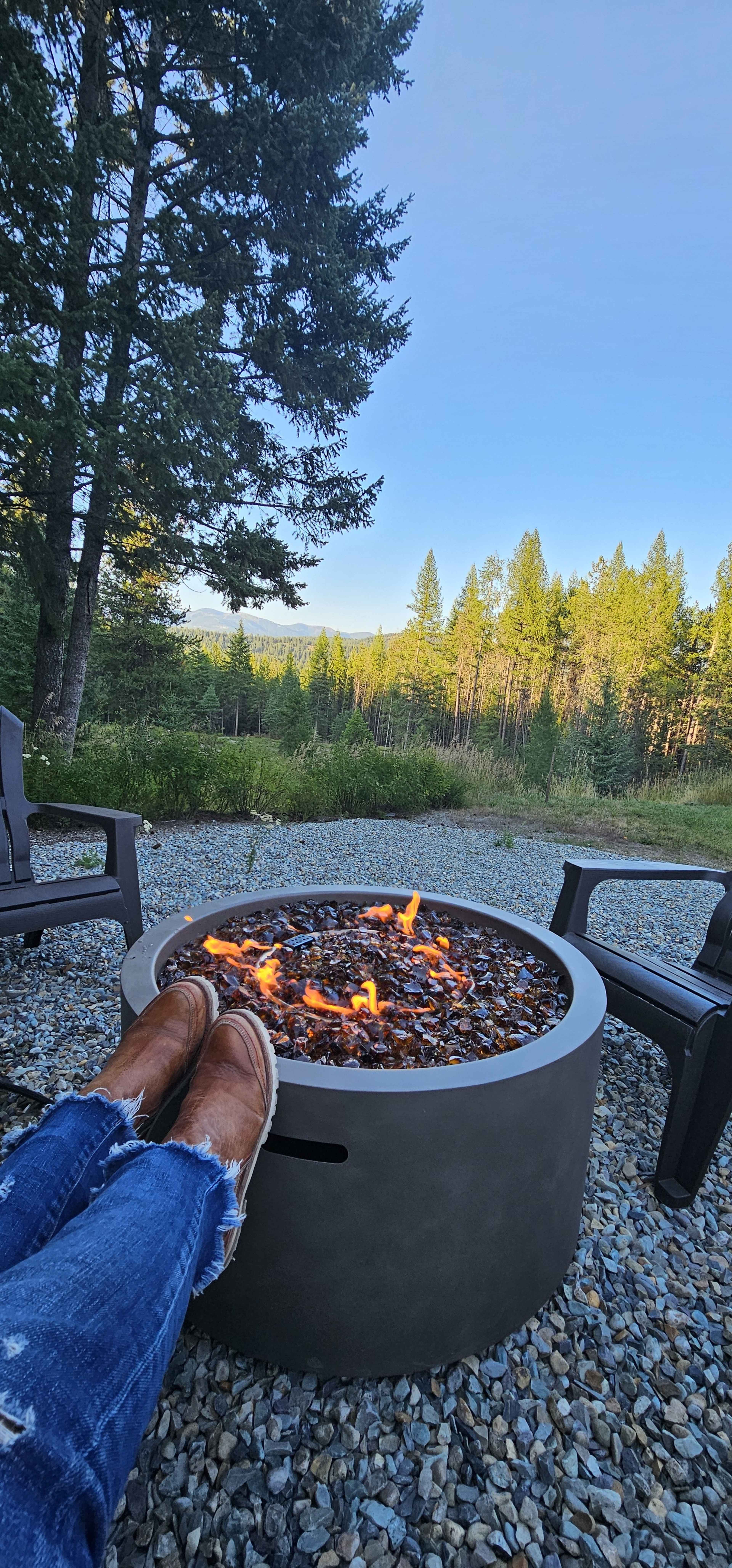 A person sits with their feet propped on the edge of a circular fire pit surrounded by gravel and wooden chairs, with a forested landscape in the background.