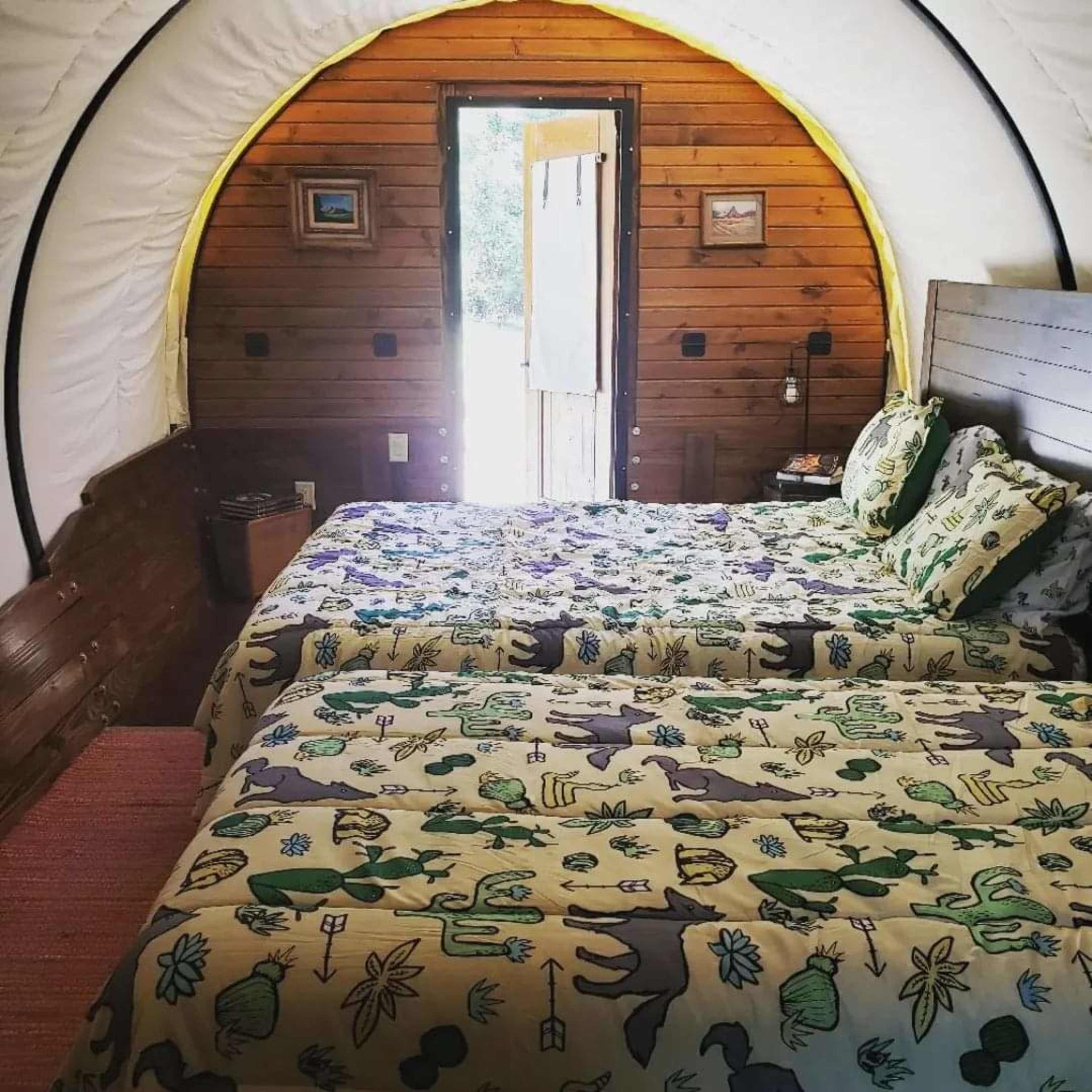 The image depicts a cozy interior of a wooden wagon-style room with two beds featuring patterned bedding and a door leading outside.