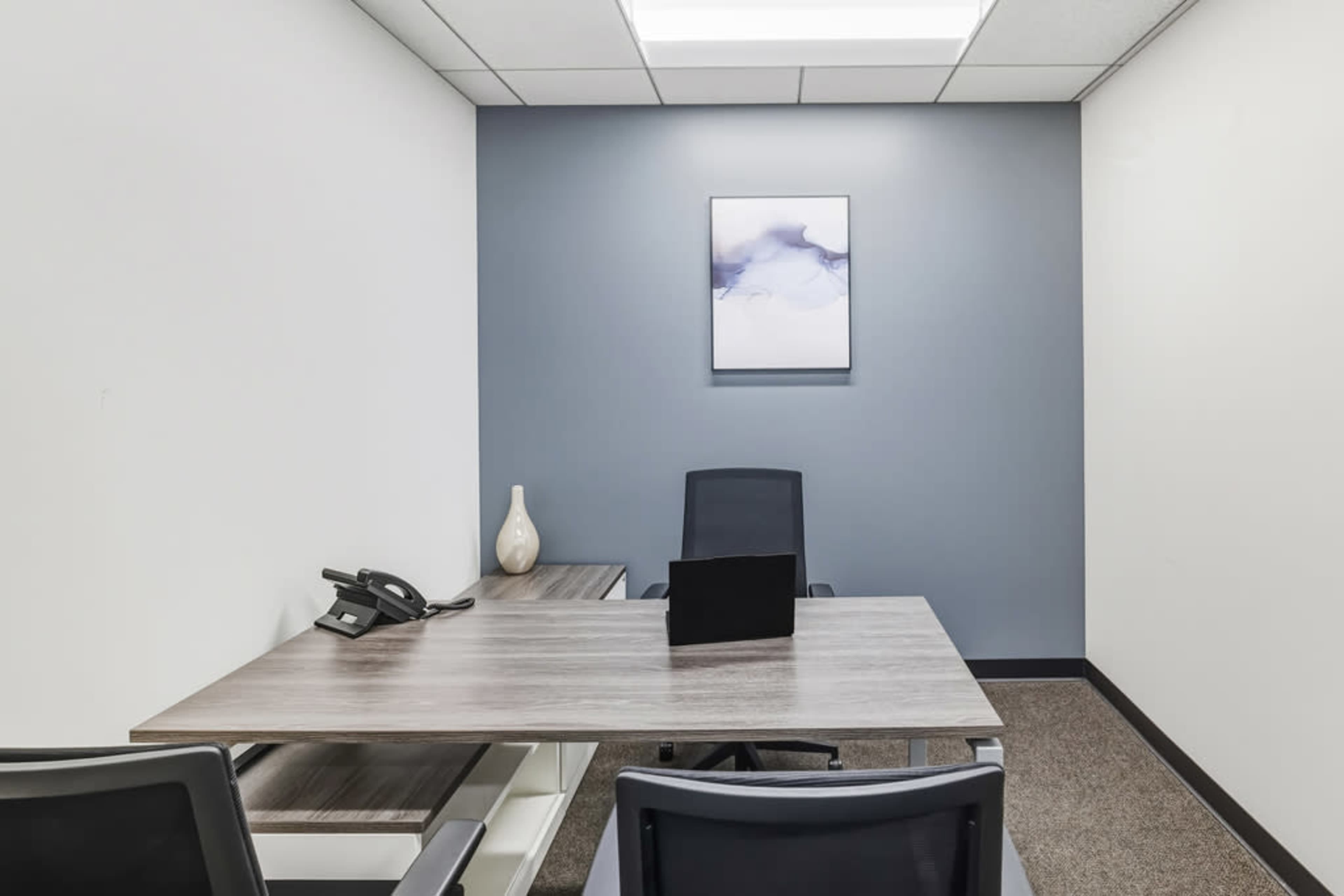 The image shows a small office space with a desk, a black chair, a telephone, and a decorative vase against a blue accent wall.