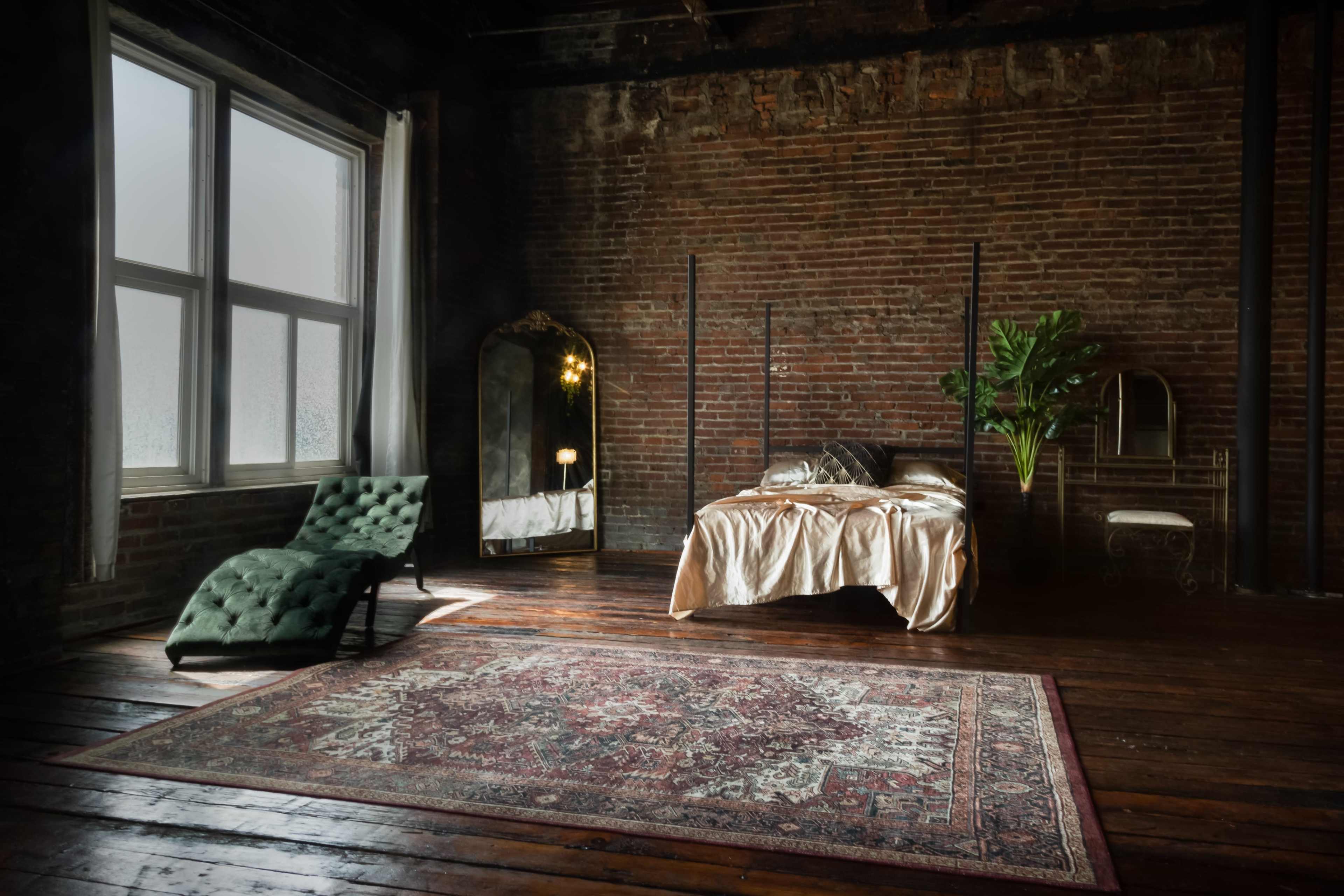 A bedroom with a vintage bed, a large mirror, a green lounge chair, and a potted plant in a room with exposed brick walls and wooden flooring.
