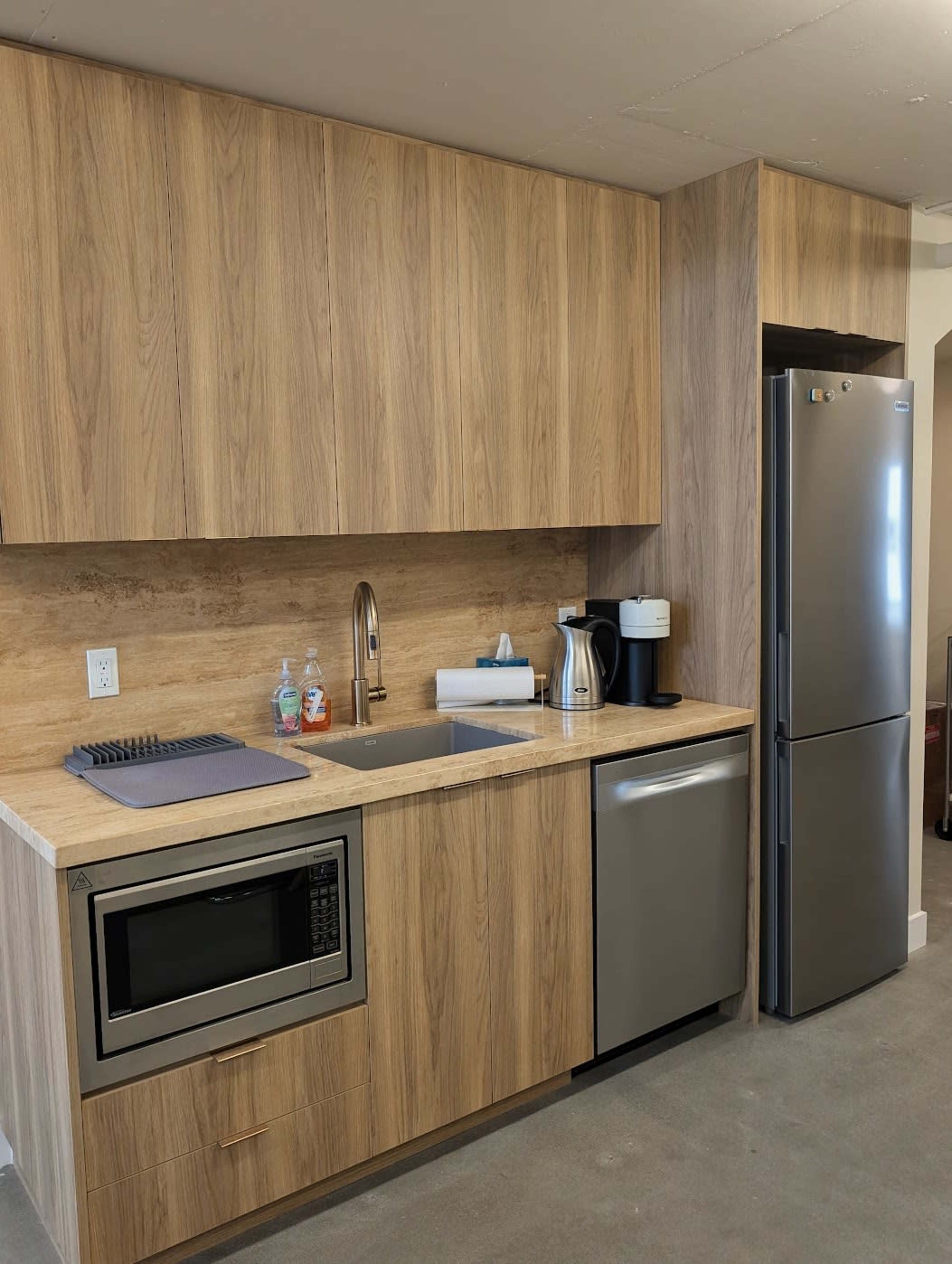The image shows a modern kitchen with wooden cabinetry, a sink, a microwave, a coffee maker, and a stainless steel refrigerator.