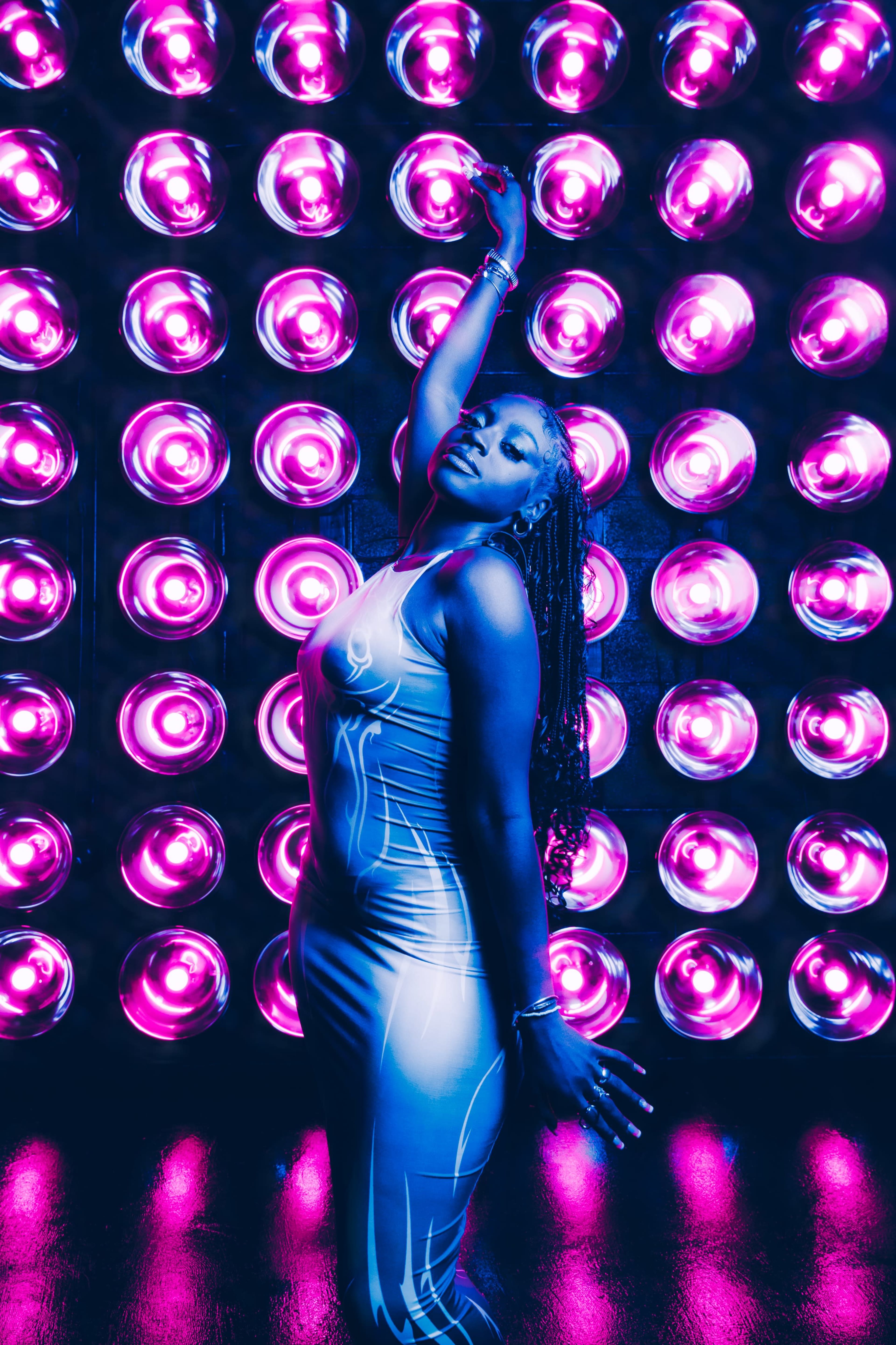 A woman poses gracefully in front of a wall of brightly lit pink and purple circular lights.