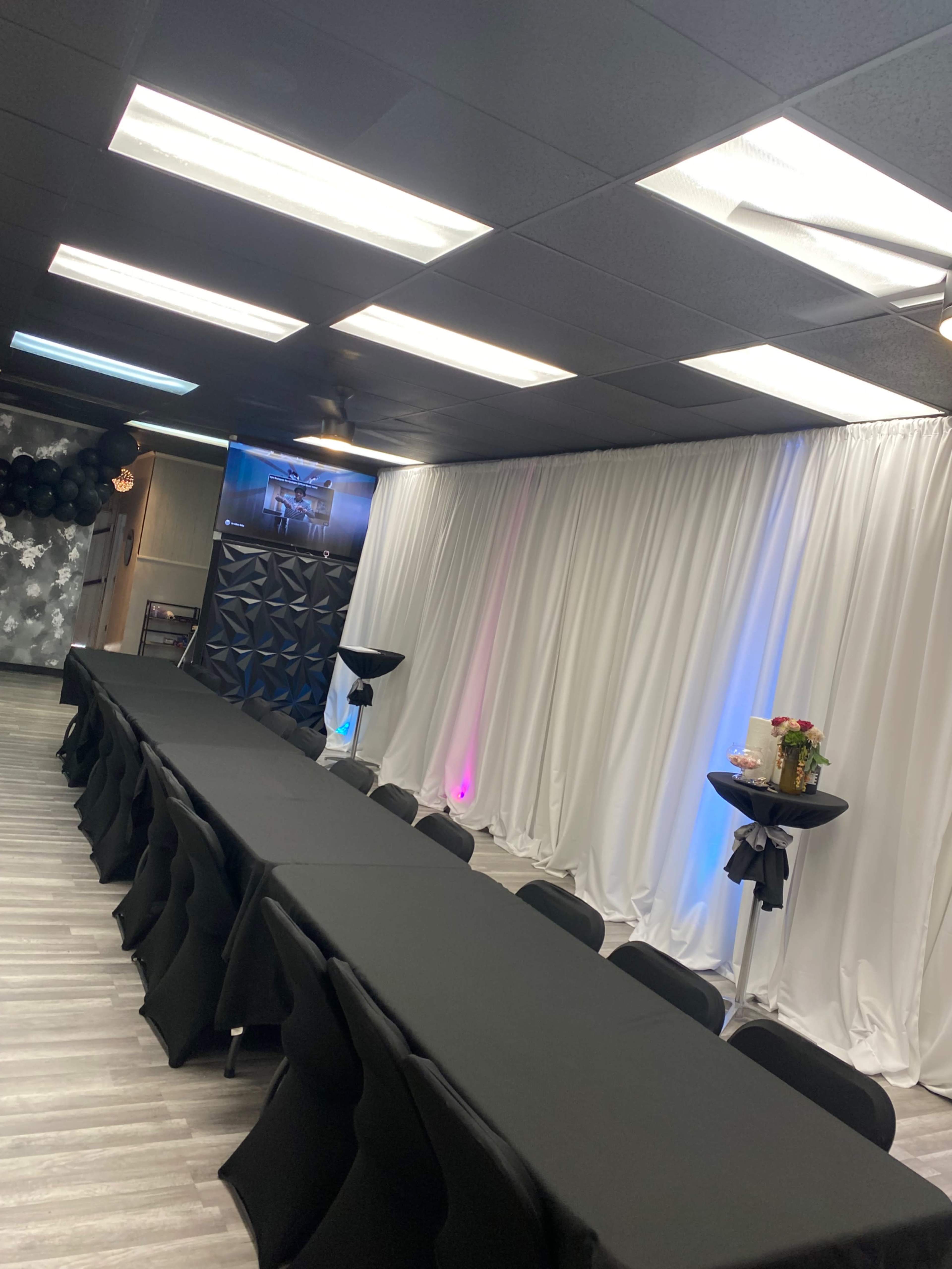 A banquet hall arranged with a long black table and tall cocktail tables, complemented by white drapes and ambient lighting.