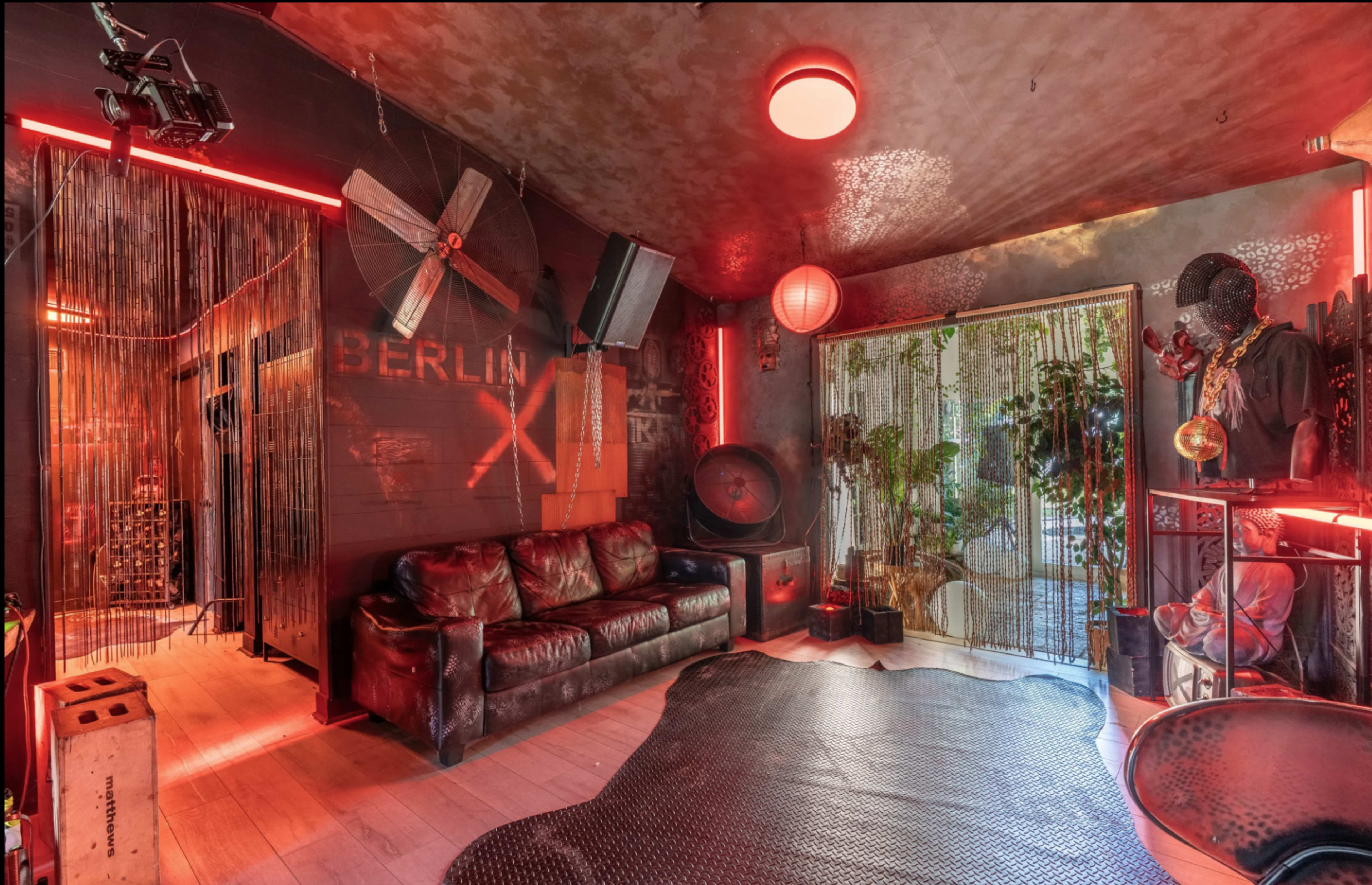 The image shows a dimly lit room with red lighting, featuring a leather sofa, decorative plants, and walls adorned with art and textures.
