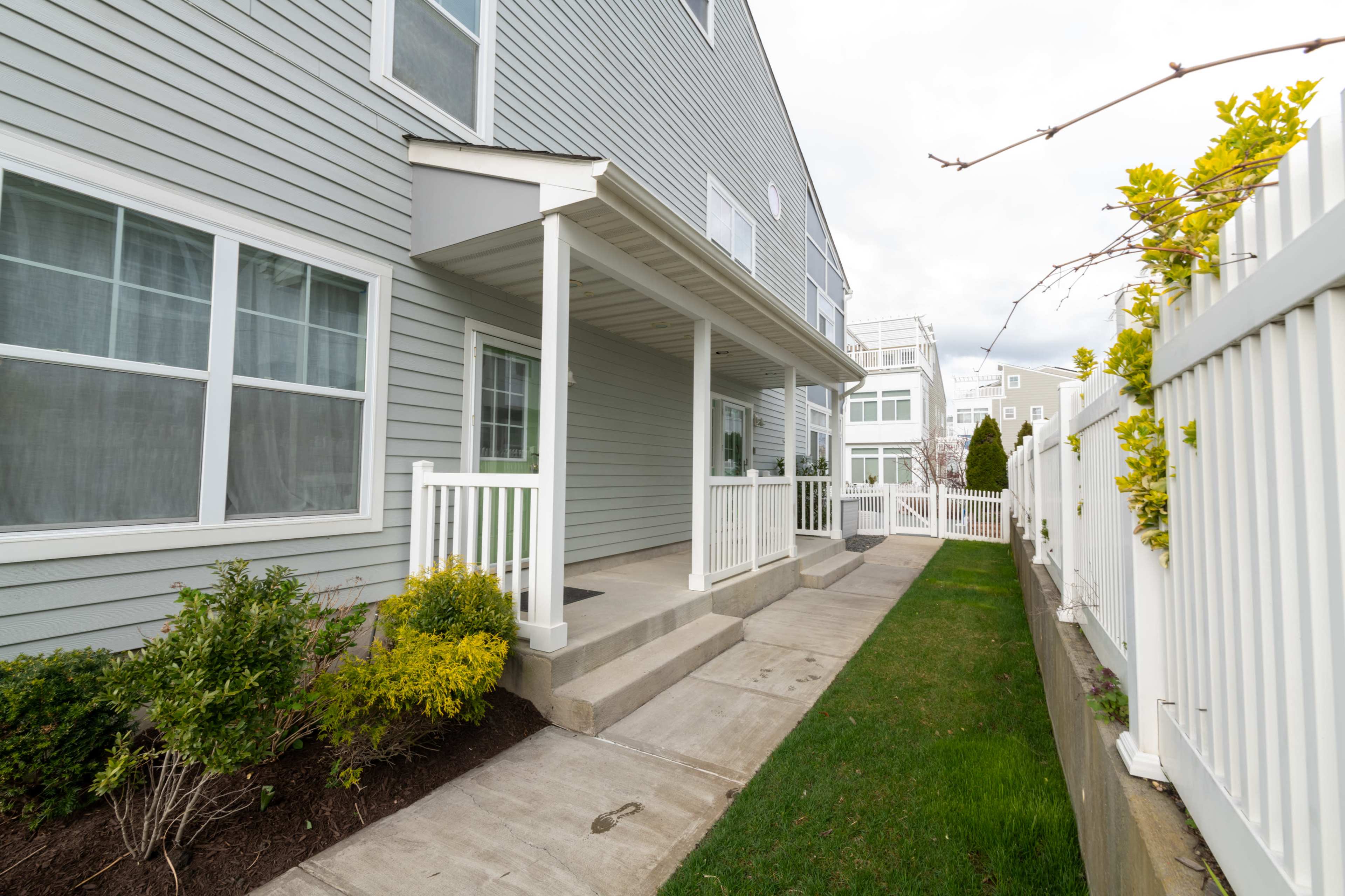 The image shows a walkway leading to a covered porch next to a gray, multi-story building, bordered by a white fence and landscaping.
