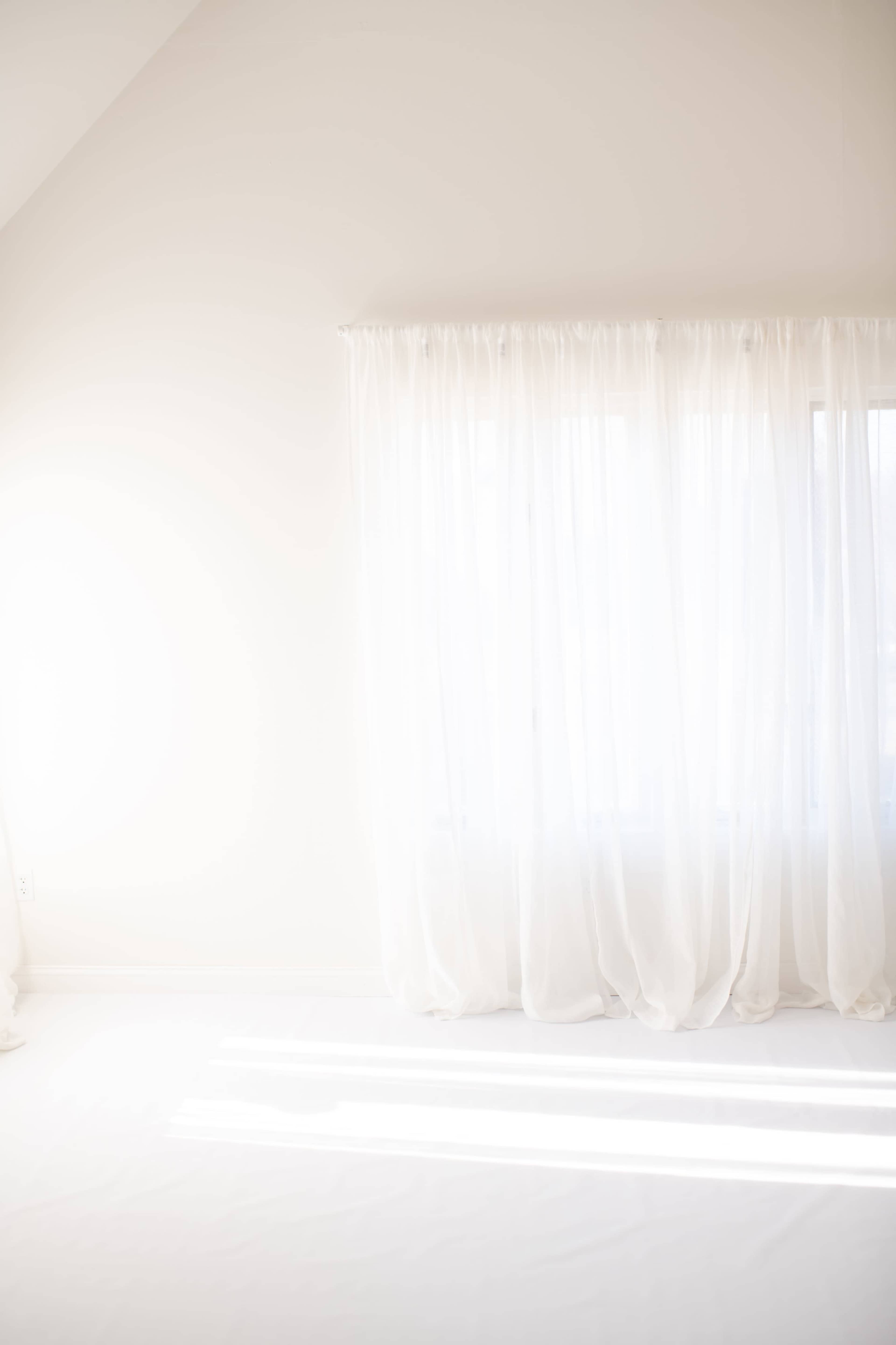 A bright, minimalistic room features sheer white curtains near a window, allowing natural light to filter in.