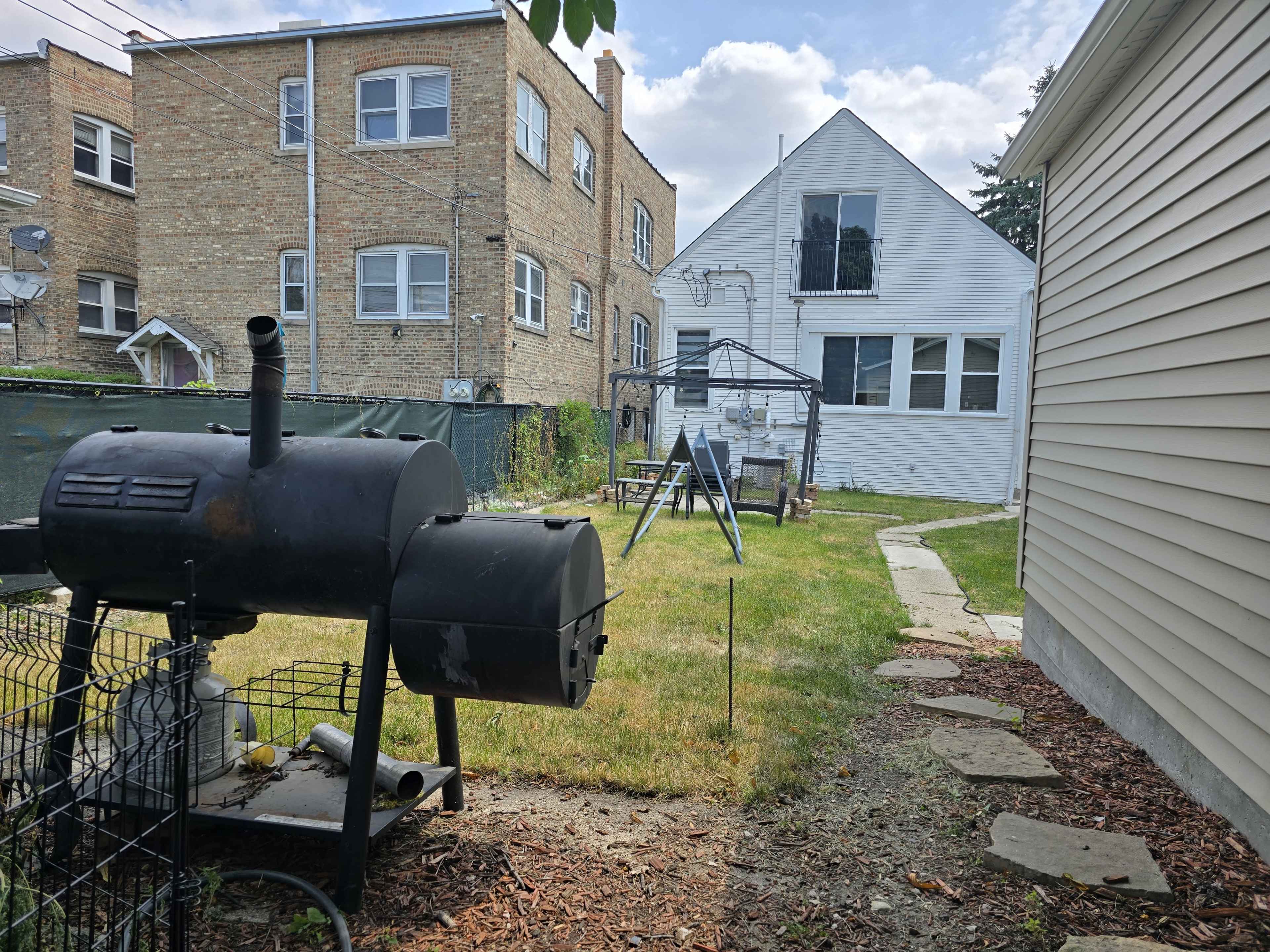 The image shows a backyard featuring a barbecue grill, a swing set, and two buildings in the background.