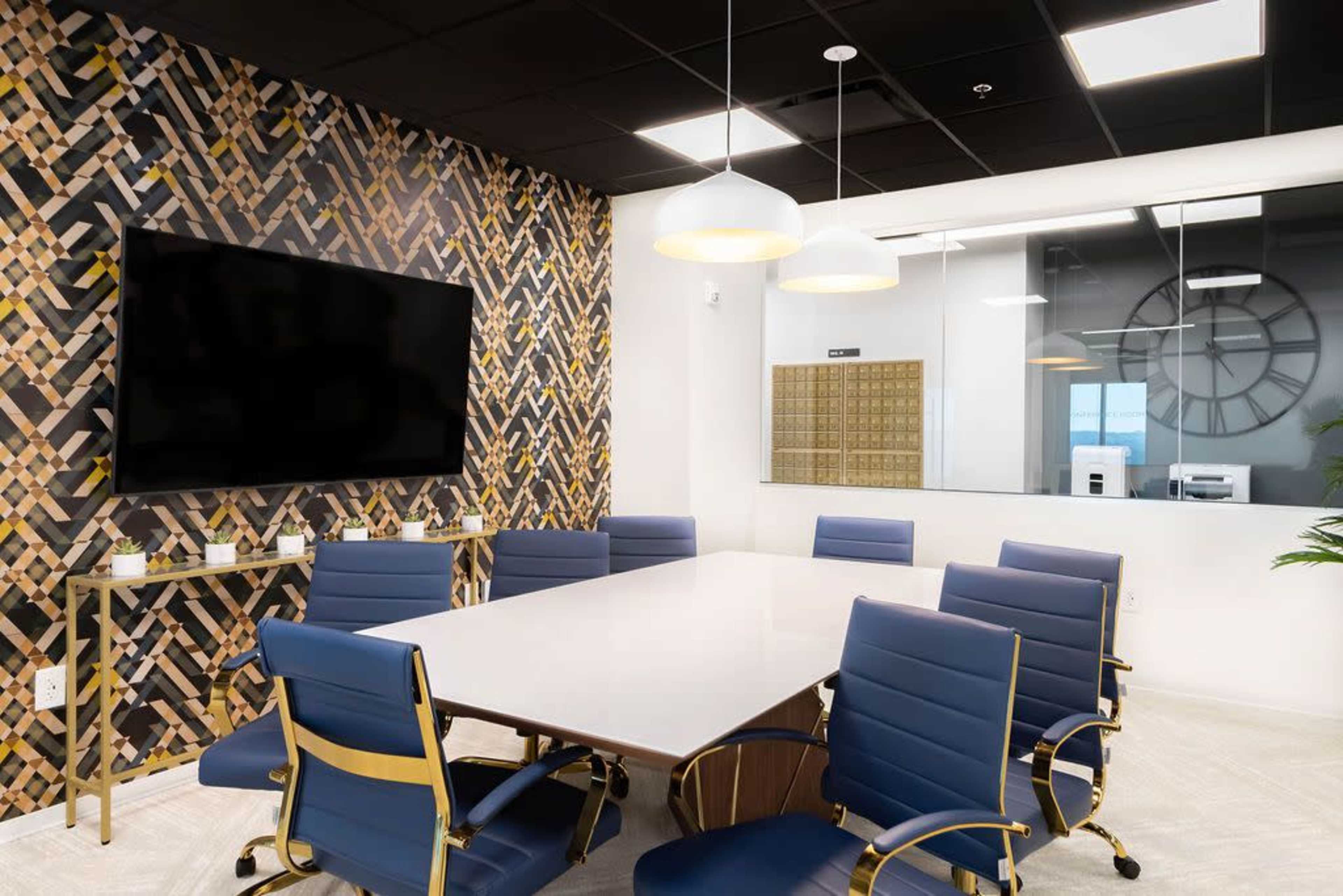 A modern conference room features a large table surrounded by blue chairs, a television on the wall, and a decorative wall with a geometric pattern.
