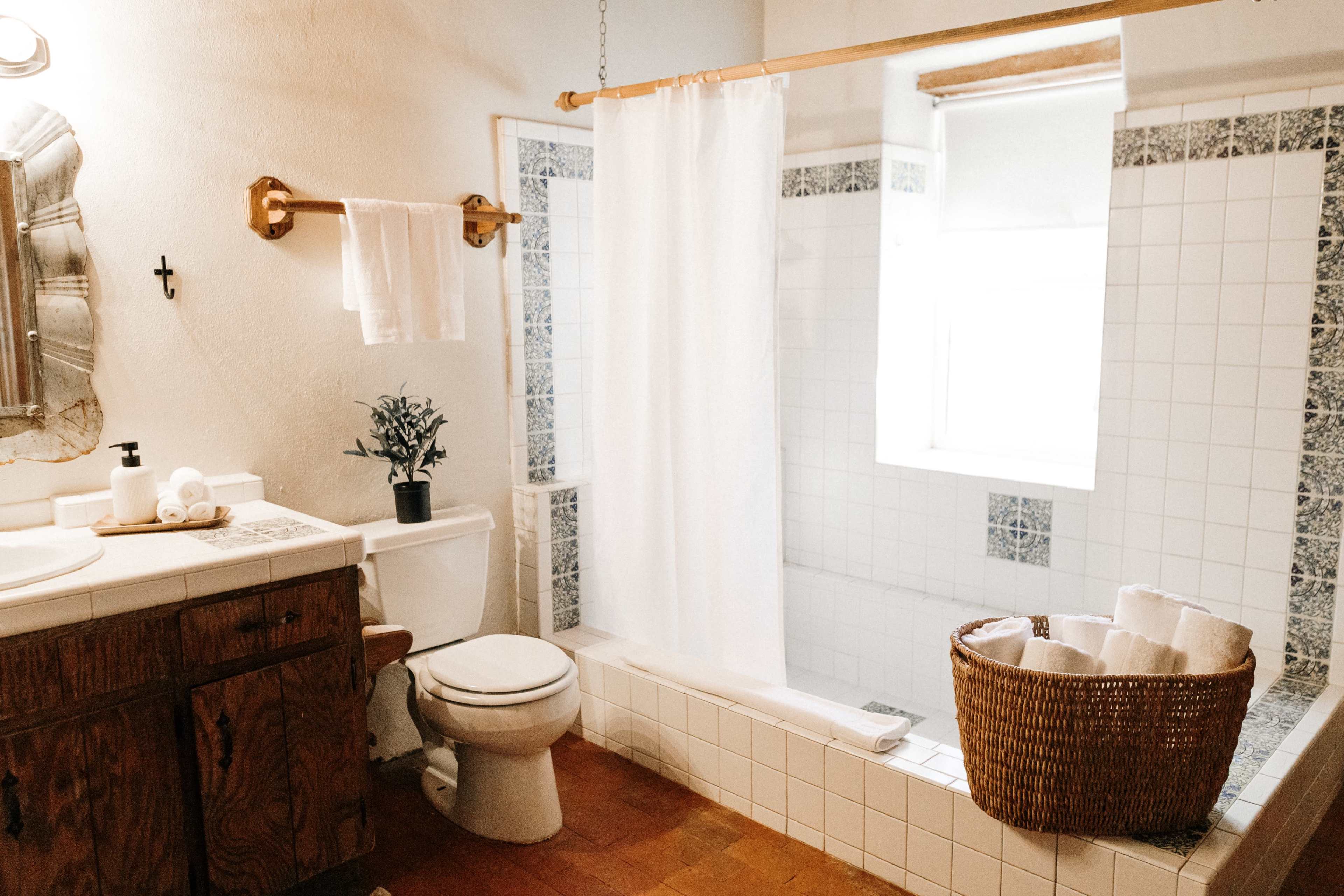 The bathroom features a shower with a white curtain, a toilet, a wooden vanity with a sink, and a wicker basket filled with rolled towels.