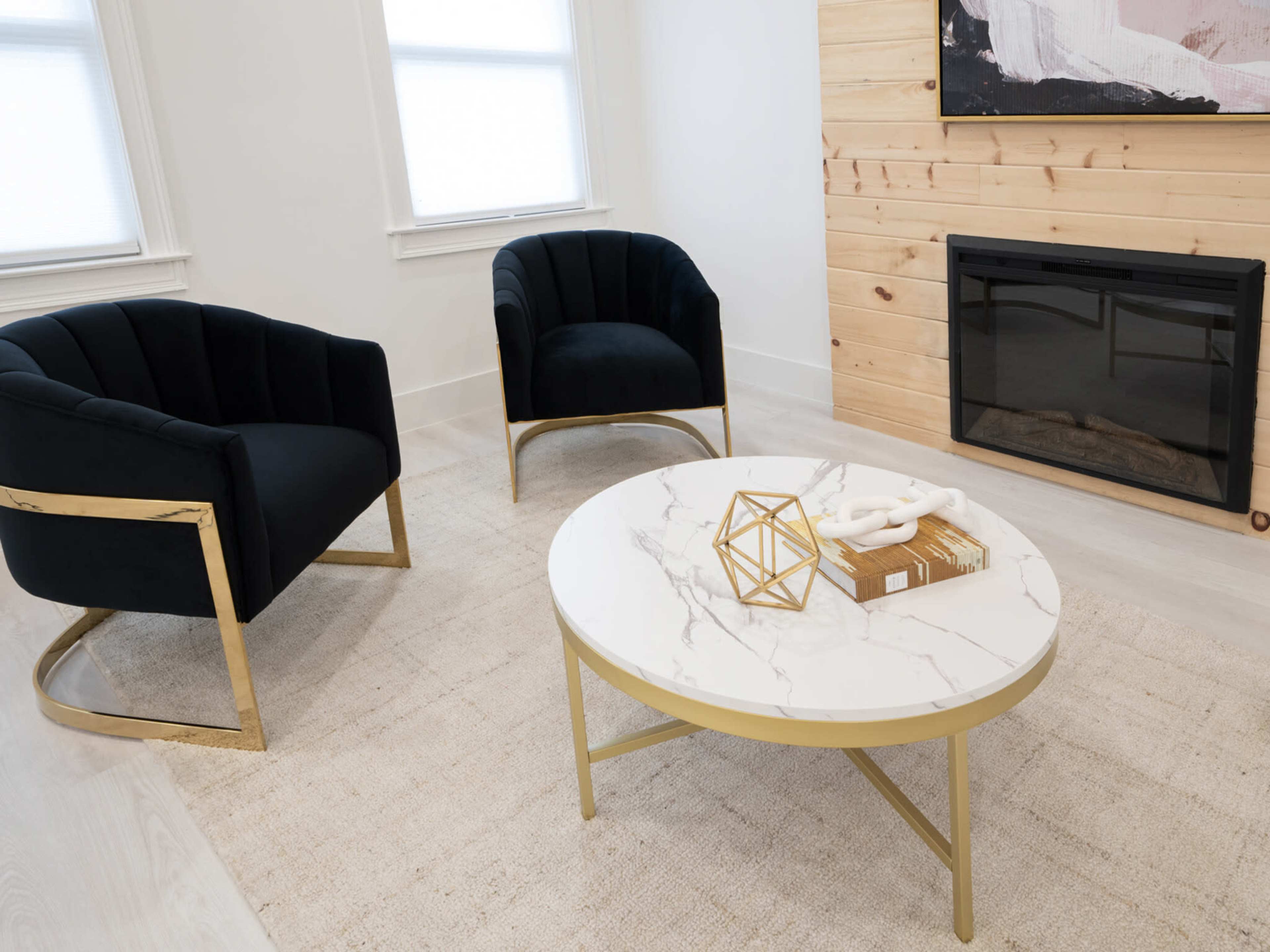 A modern living room with two black chairs, a round marble coffee table, and a fireplace against a wooden wall.