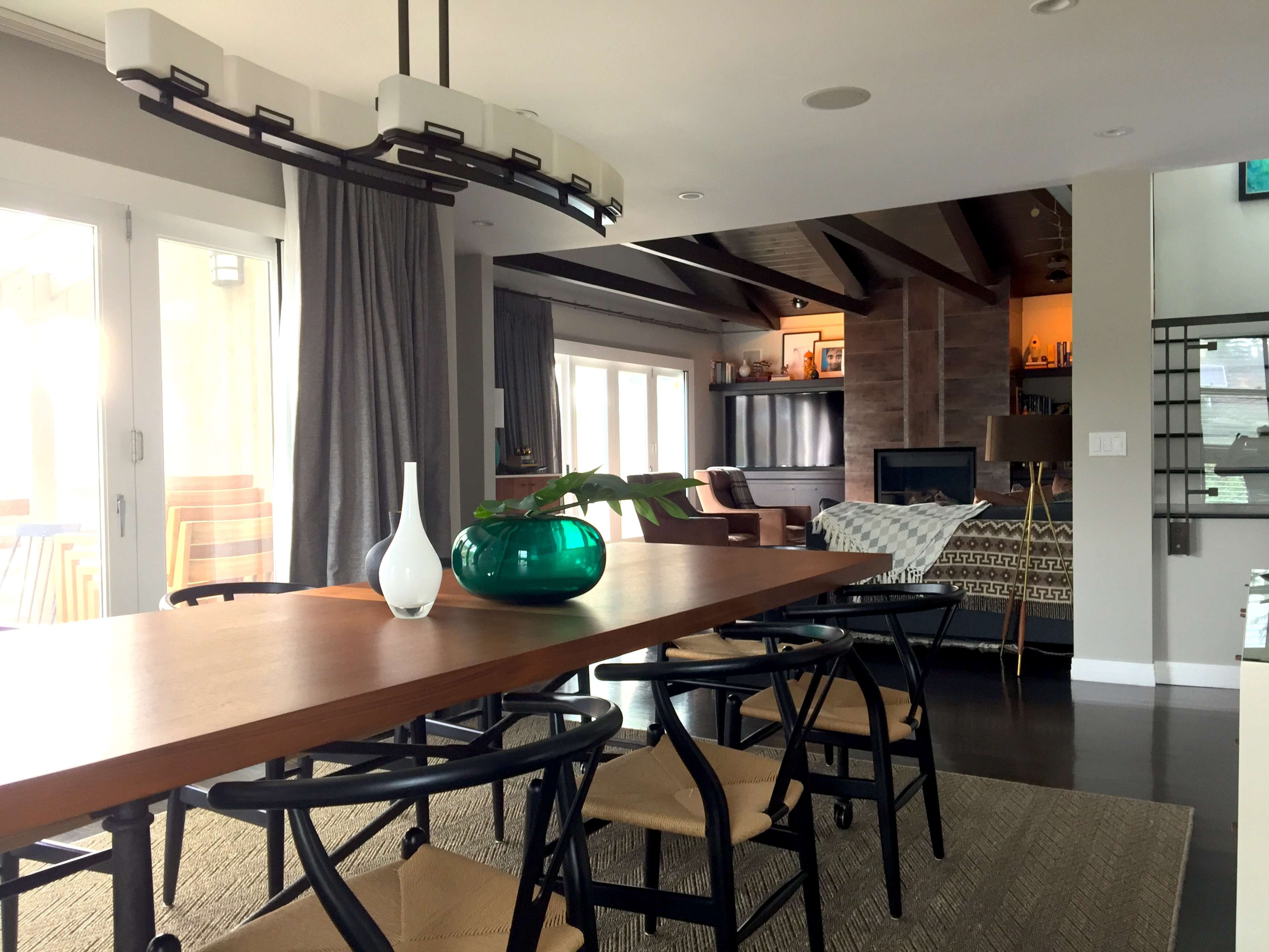 A modern dining area features a wooden table with a green glass vase and a view of a living space with a cozy couch and fireplace.