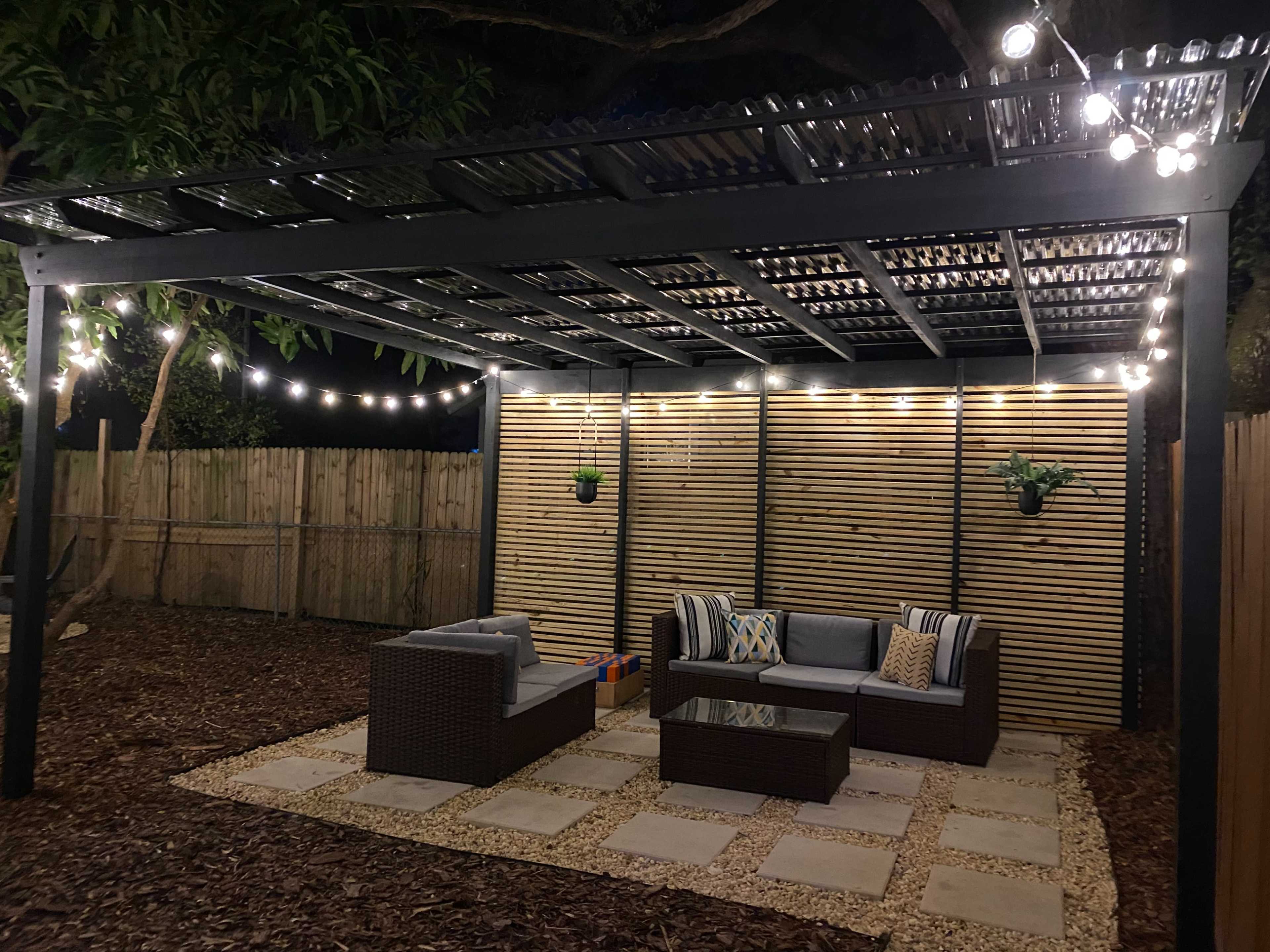 A patio area features a modern pergola with string lights, surrounded by landscaping and furnished with a seating arrangement and coffee table.