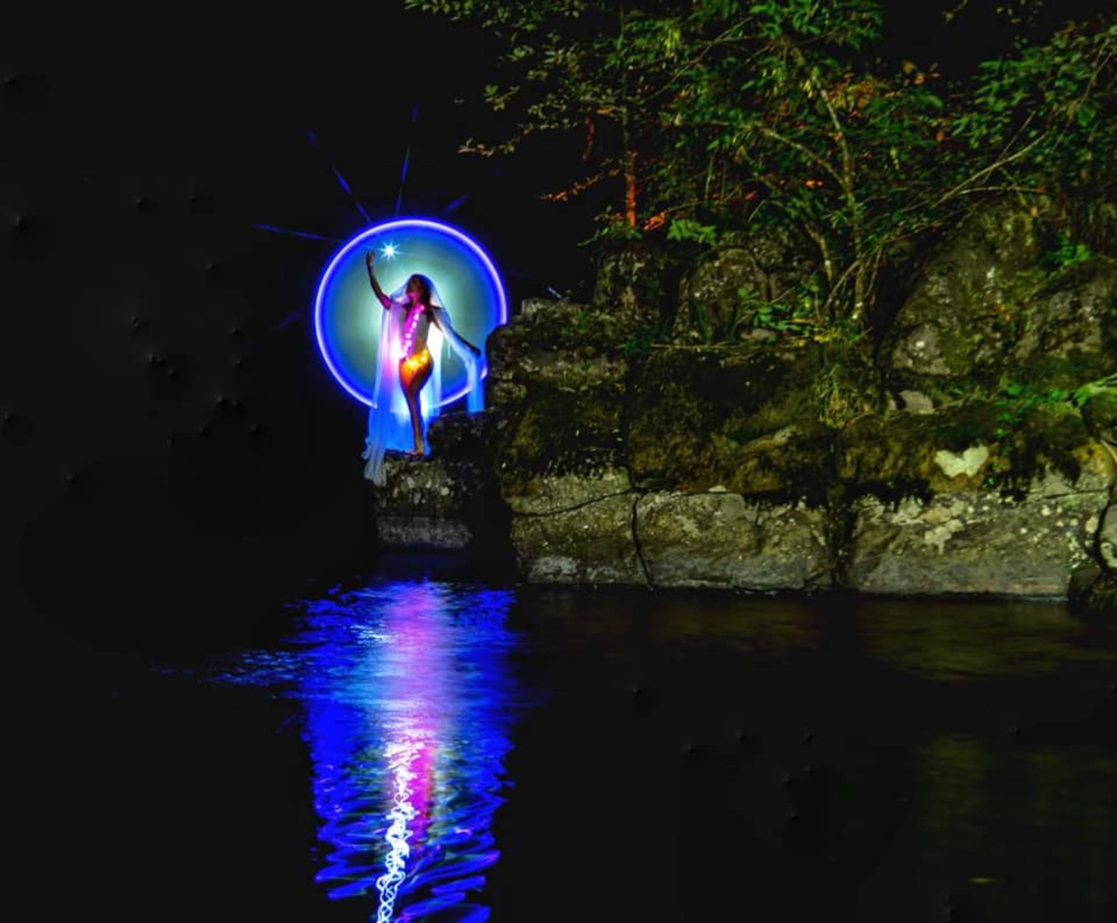 A figure illuminated by colored lights stands on a rock beside a body of water, surrounded by trees in a dark setting.