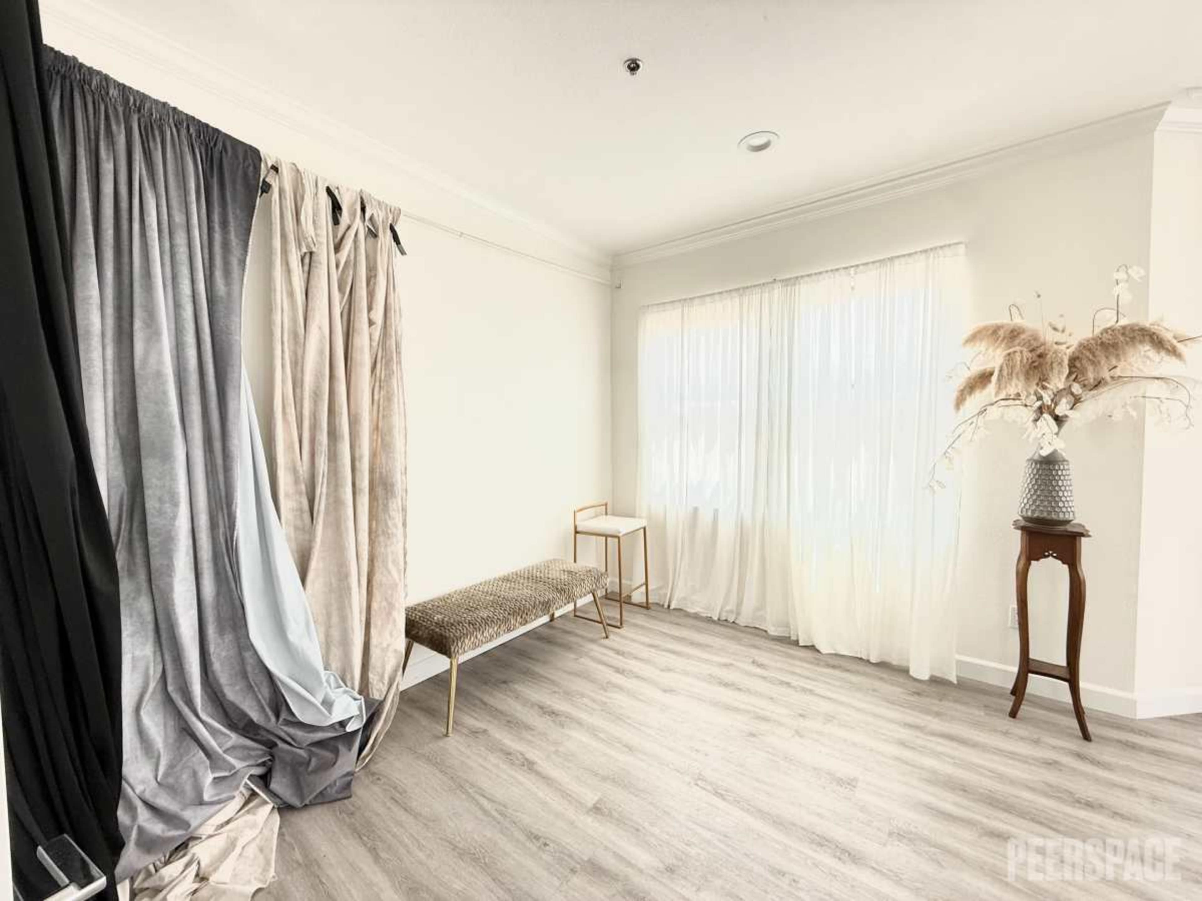 The image shows a bright room with light-colored walls, a large window draped with sheer curtains, a decorative bench, and standing drapes in various shades.