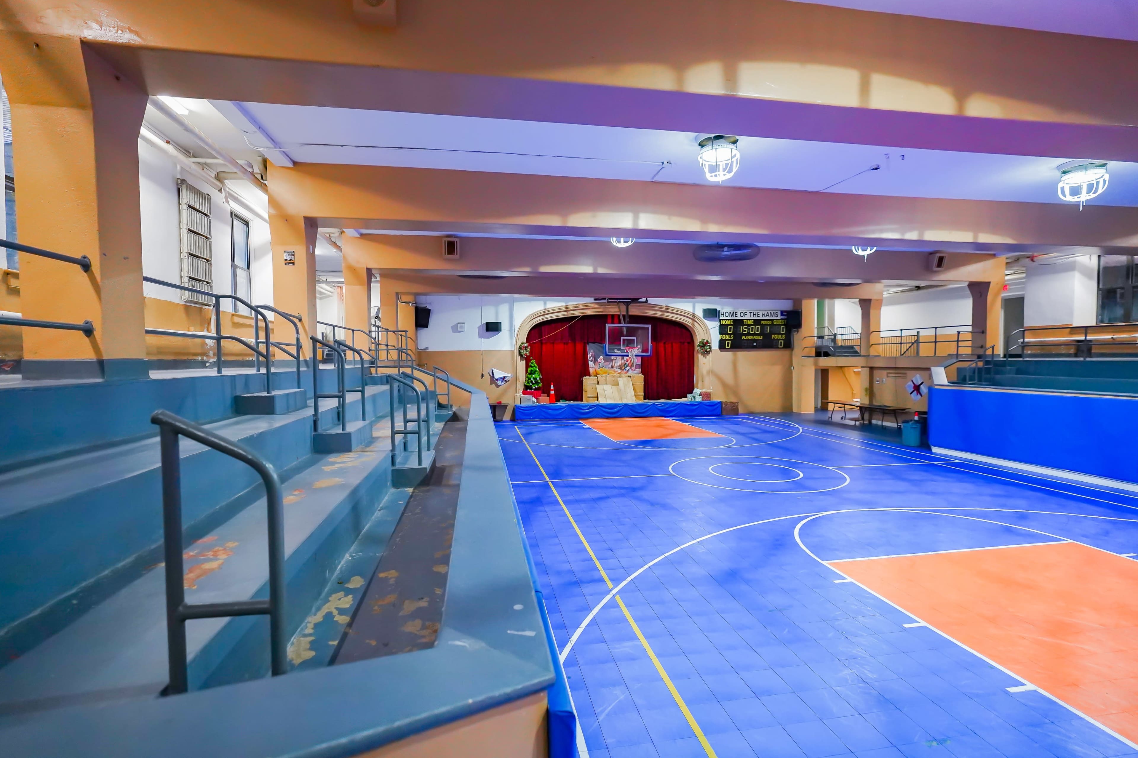 A gymnasium with a blue and orange basketball court and bleachers, featuring a stage draped in red at the far end.