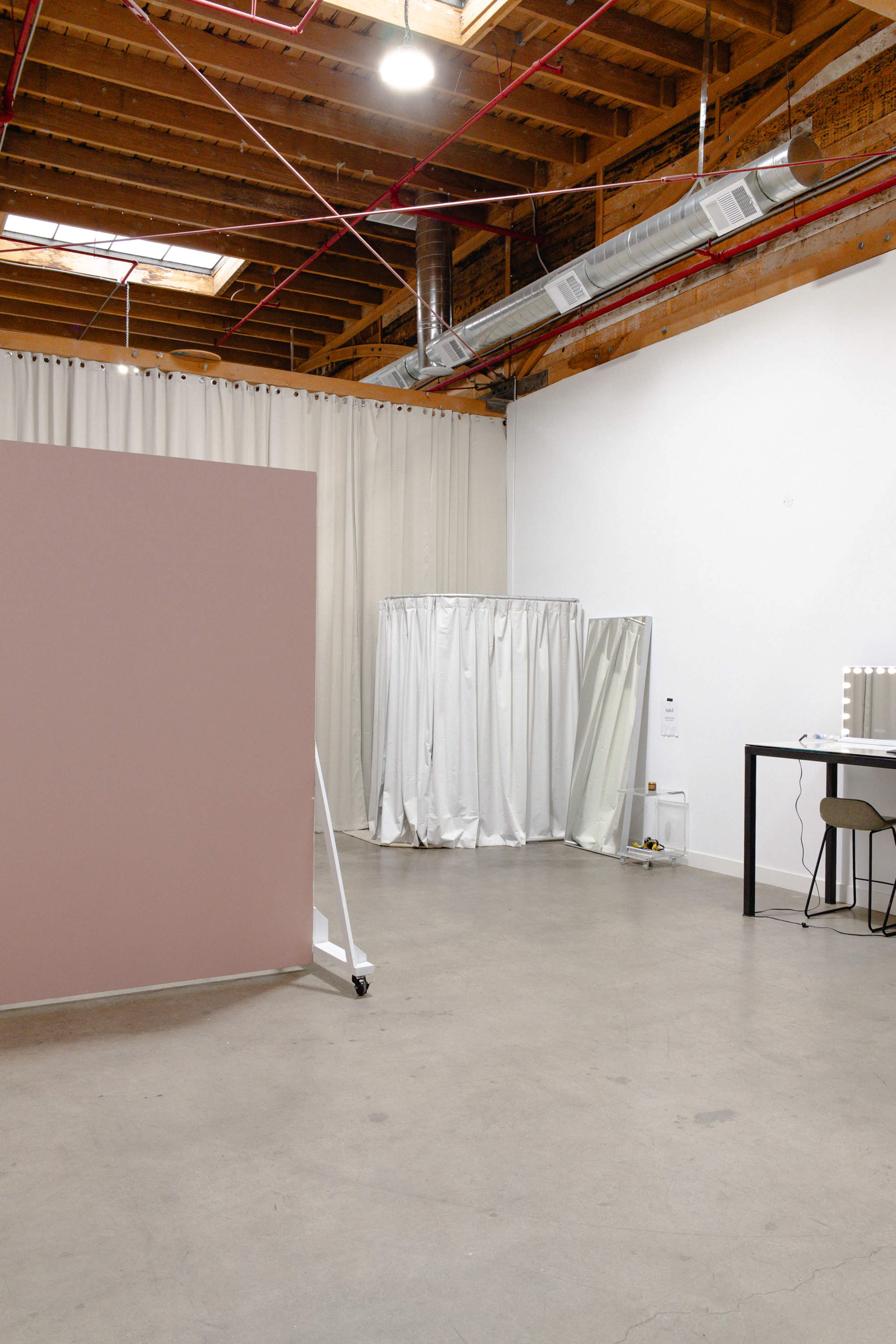 The image shows a spacious interior with a pink partition, white curtains, a mirror, and a table with a chair against a wall.