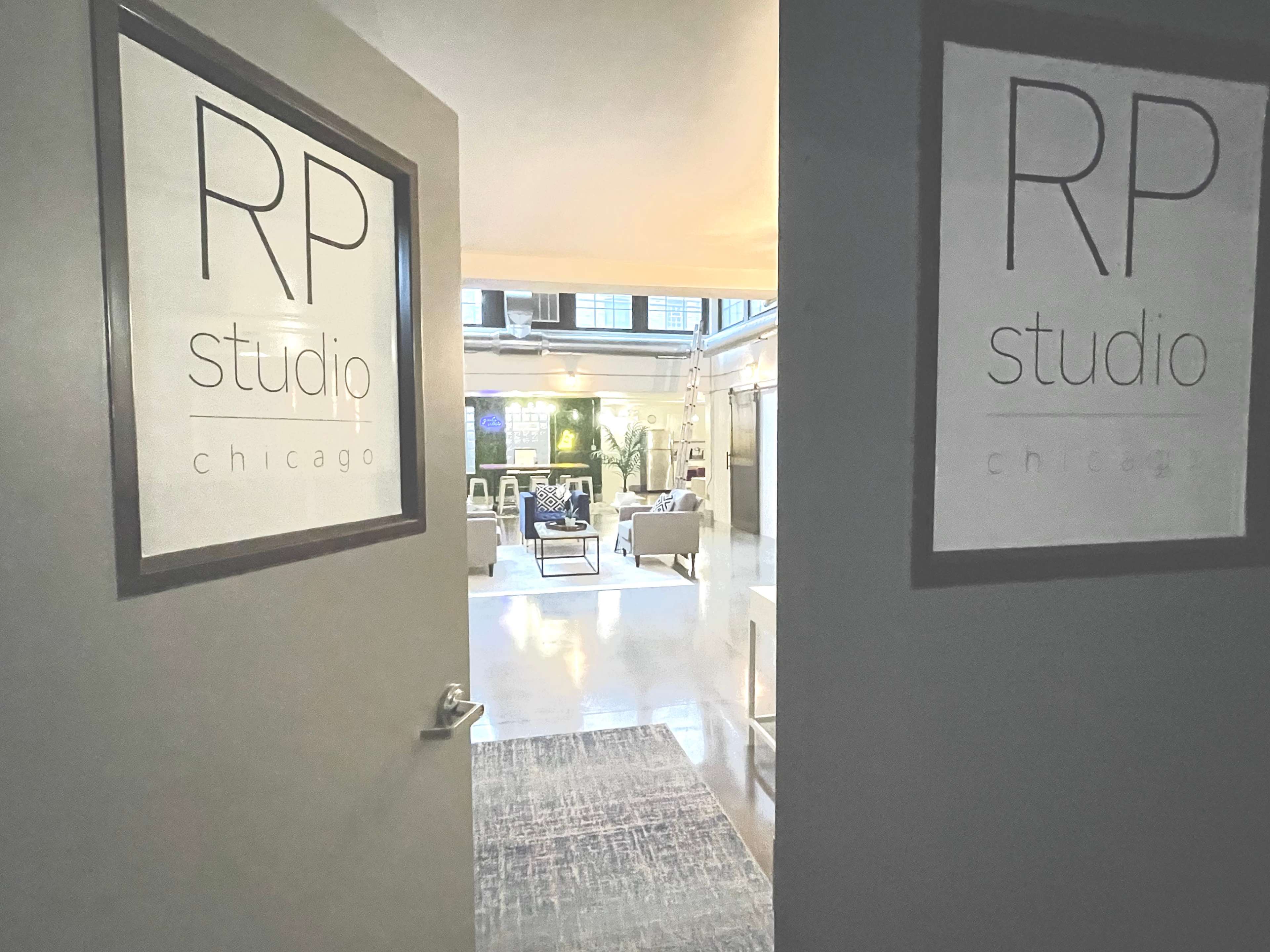 The image shows an open doorway leading into a modern studio space with a sitting area that includes sofas and large windows.