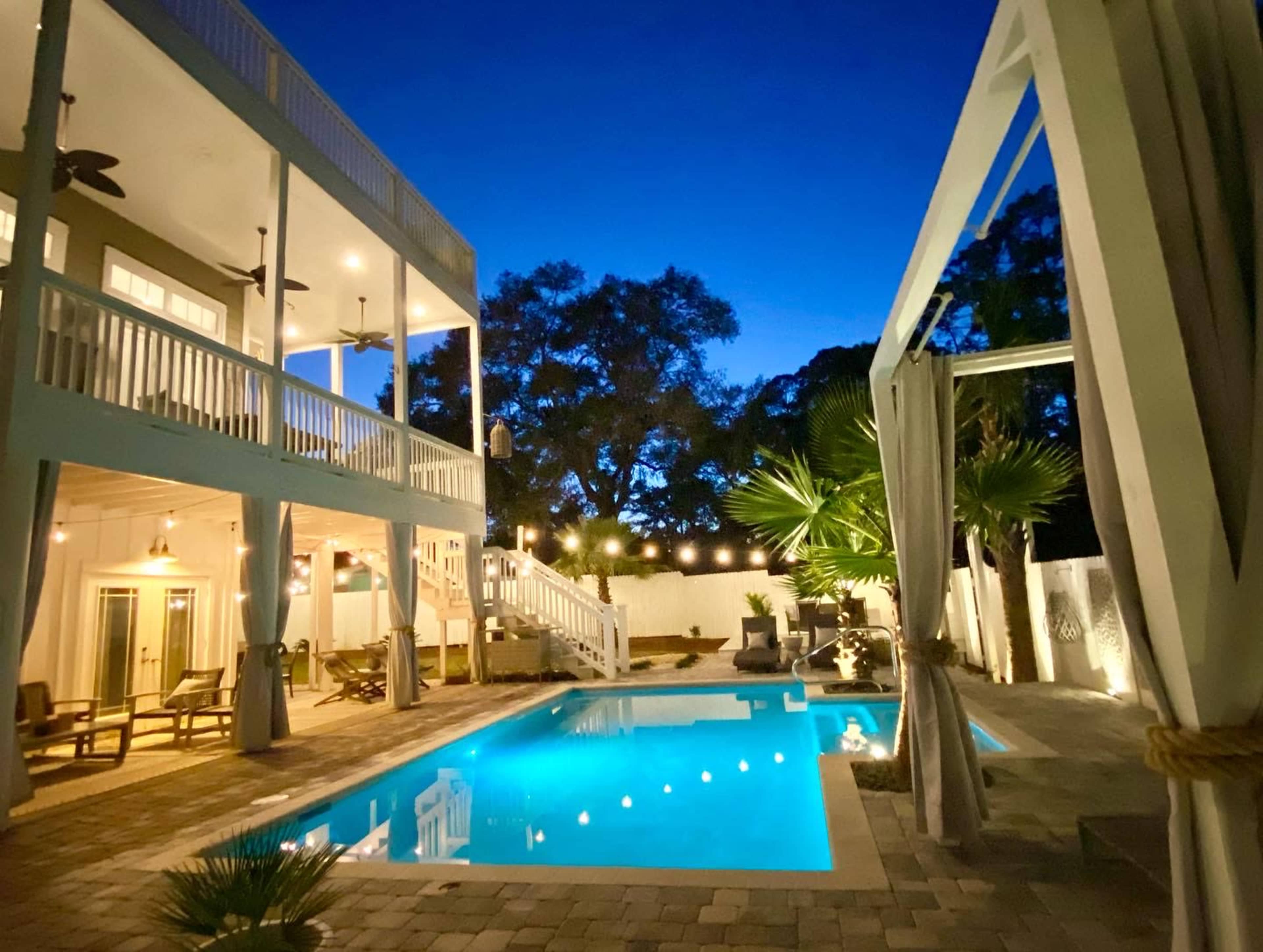 A modern house with a swimming pool lit up at night, surrounded by palm trees and outdoor seating areas.
