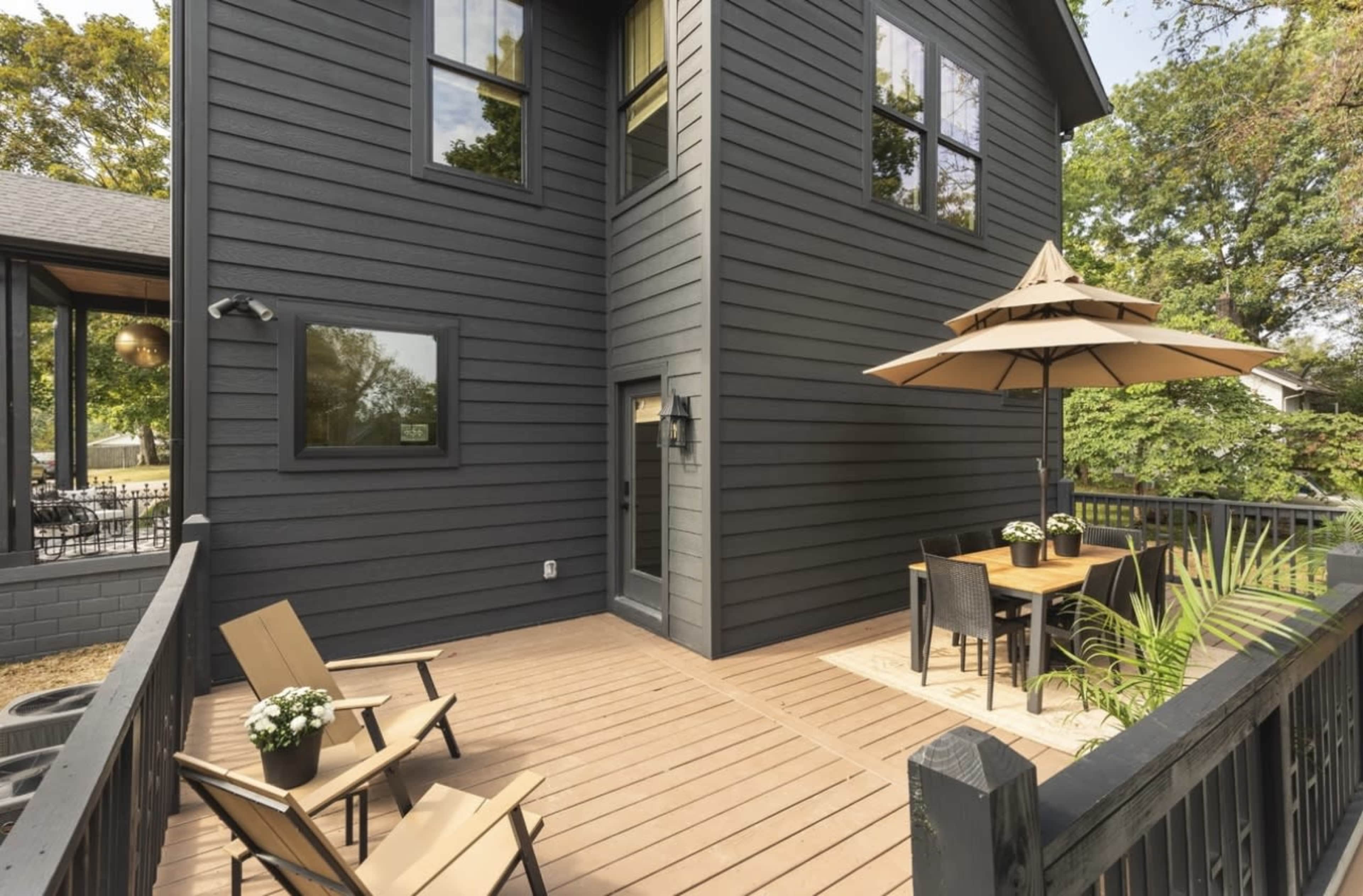 The image shows a two-story house with a dark exterior, featuring a wooden deck that has outdoor furniture and an umbrella.