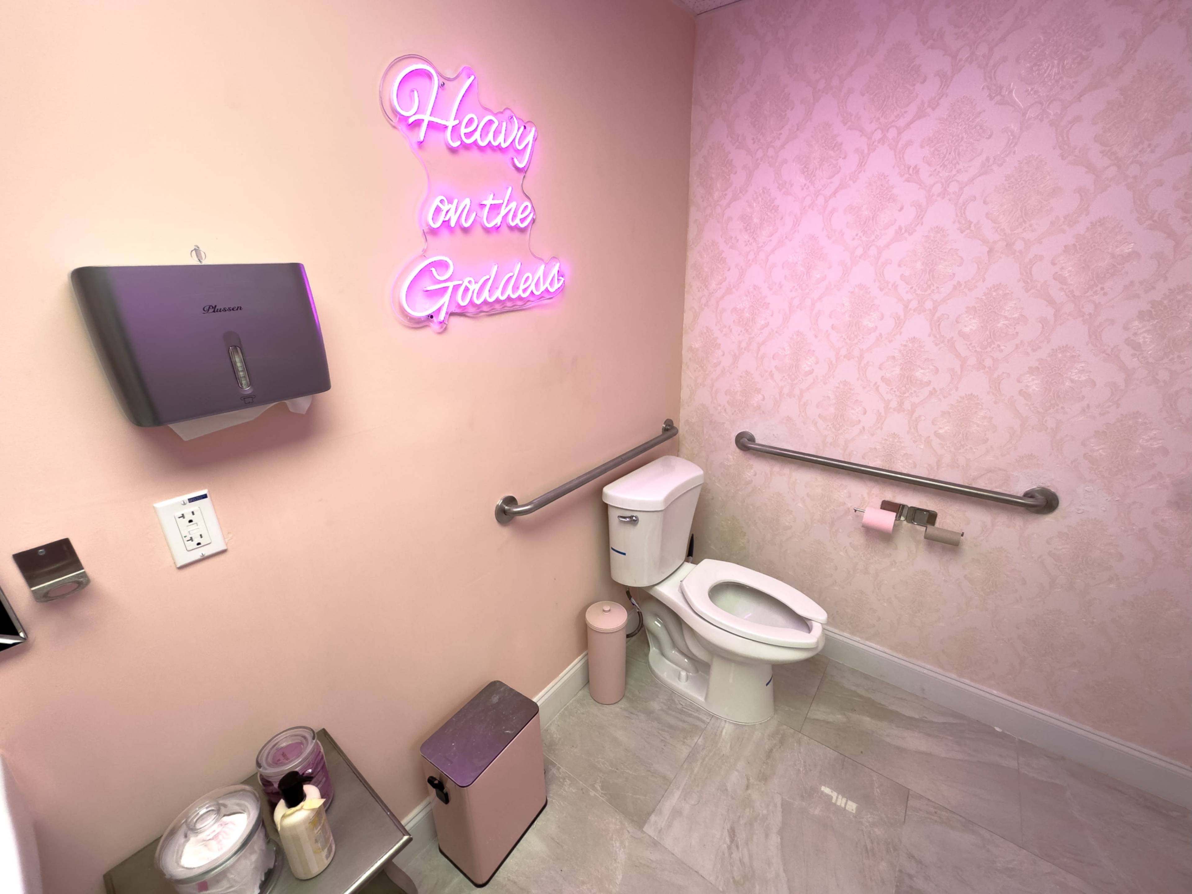 A restroom features a toilet, hand dryer, and a neon sign that reads "Heavy on the Goddess" against a pink patterned wall.