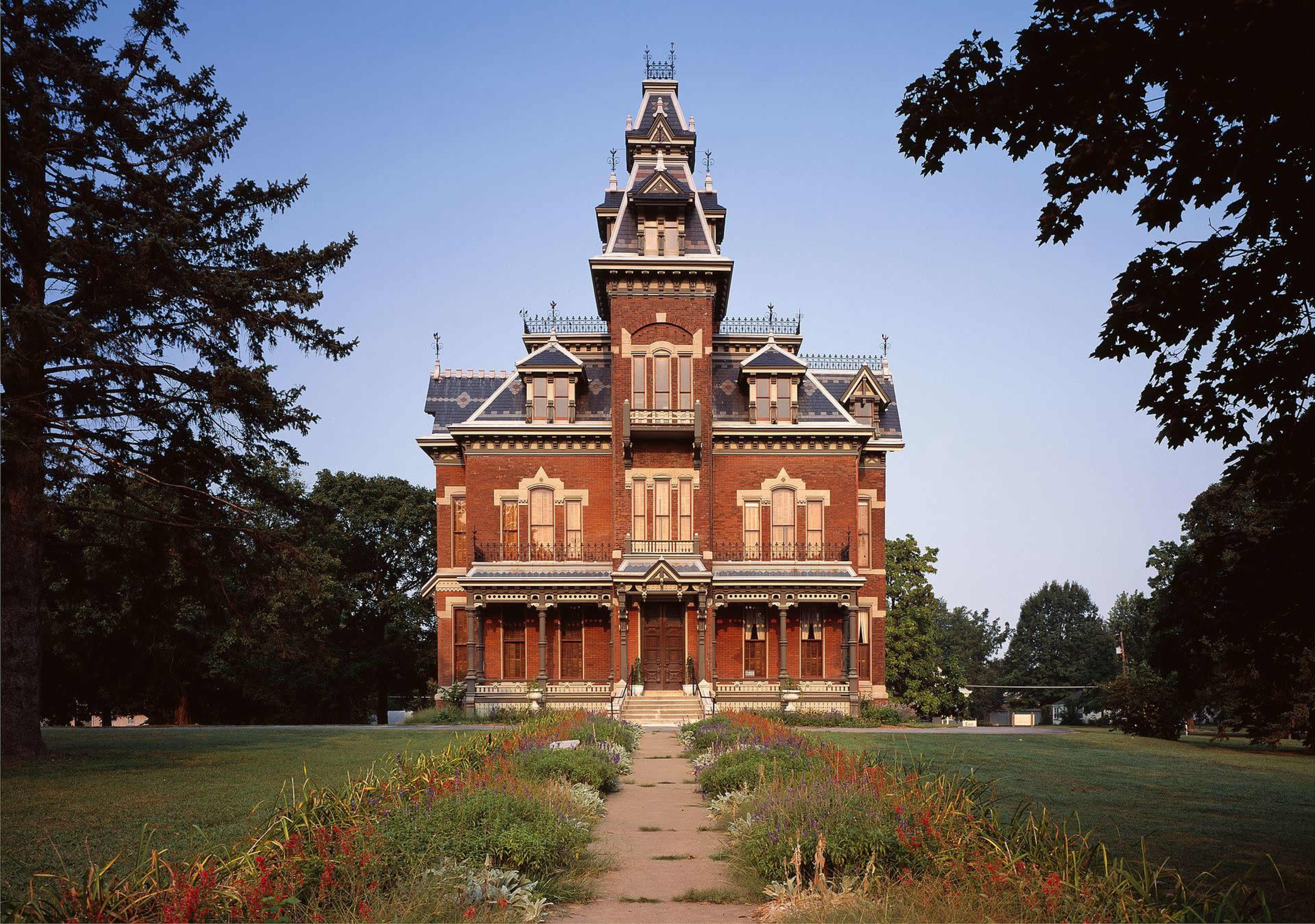 A large Victorian-style mansion with intricate architectural details stands prominently at the end of a pathway, surrounded by landscaped gardens.