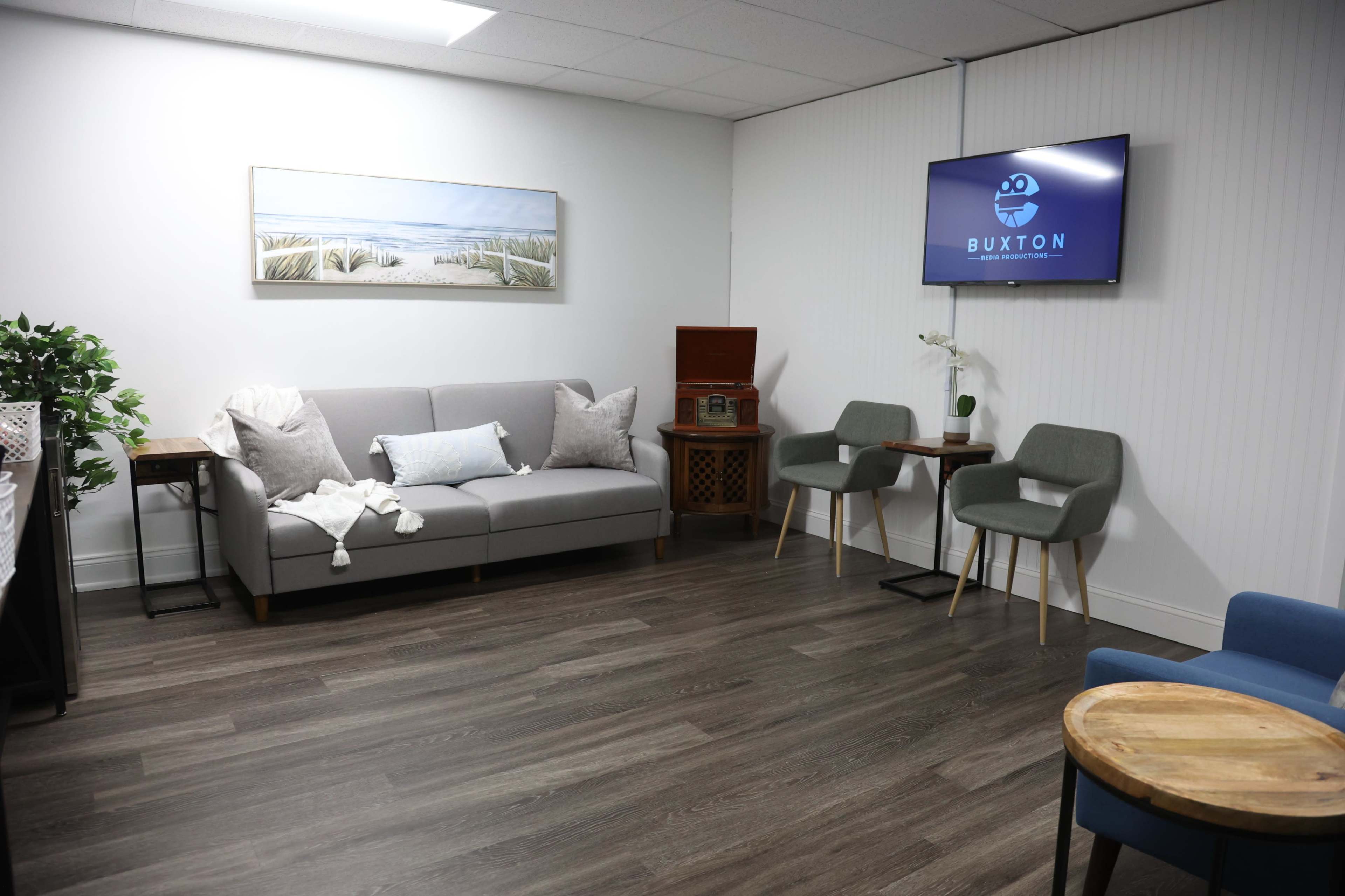 The image shows a modern waiting room featuring a sofa, two chairs, a wooden side table, an antique cabinet, and a television displaying the name "Buxton."