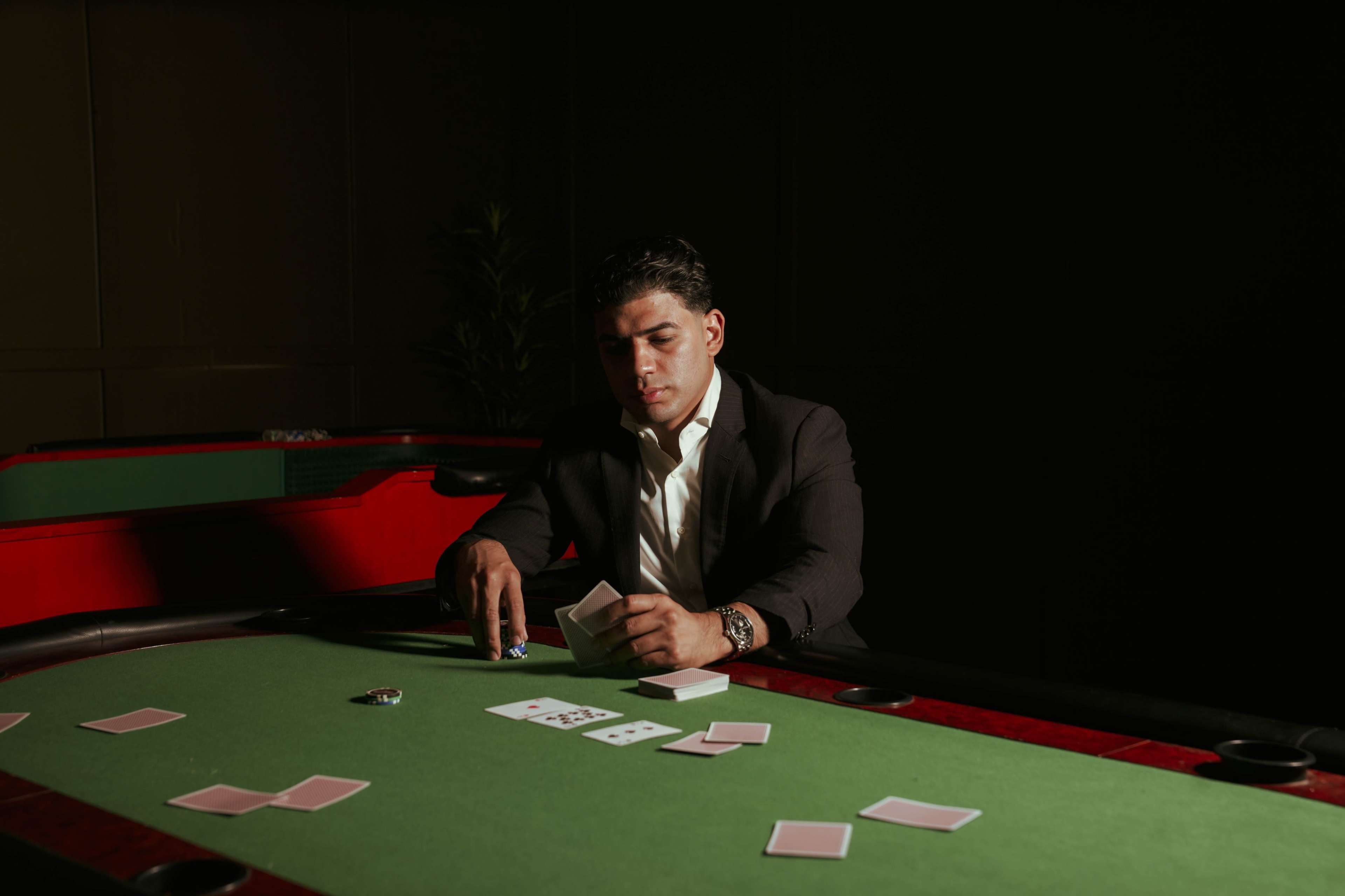 A man in a black suit sits at a green felt poker table, examining a set of playing cards with a focused expression.