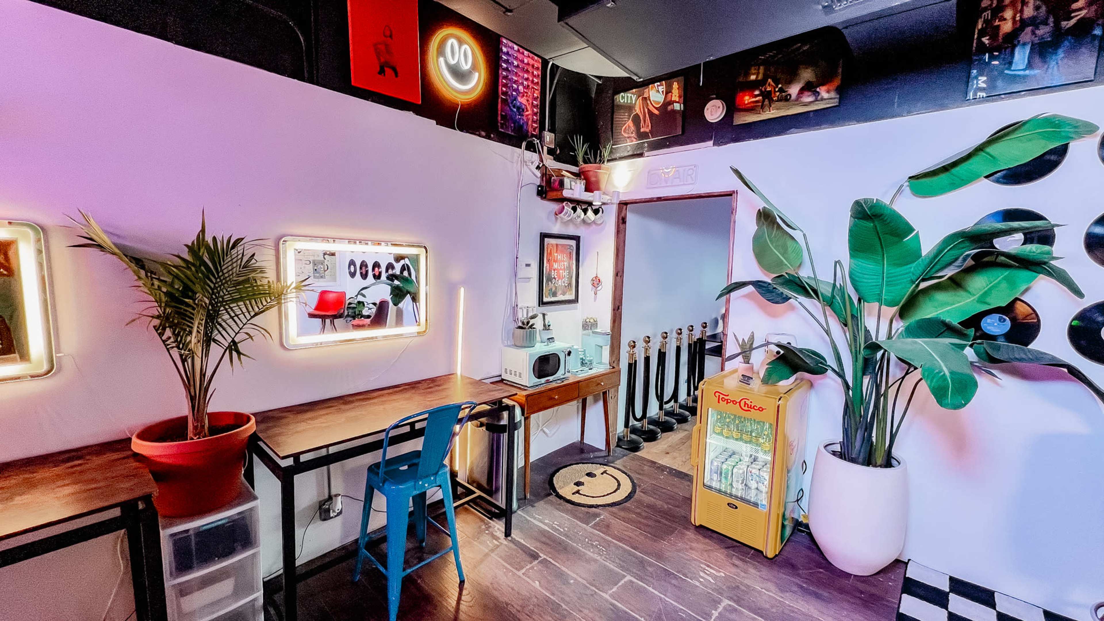 The image shows a cozy room with a small kitchen area featuring a microwave, a fridge, a wooden desk with a blue chair, large potted plants, and decorative wall items including vinyl records and neon signs.