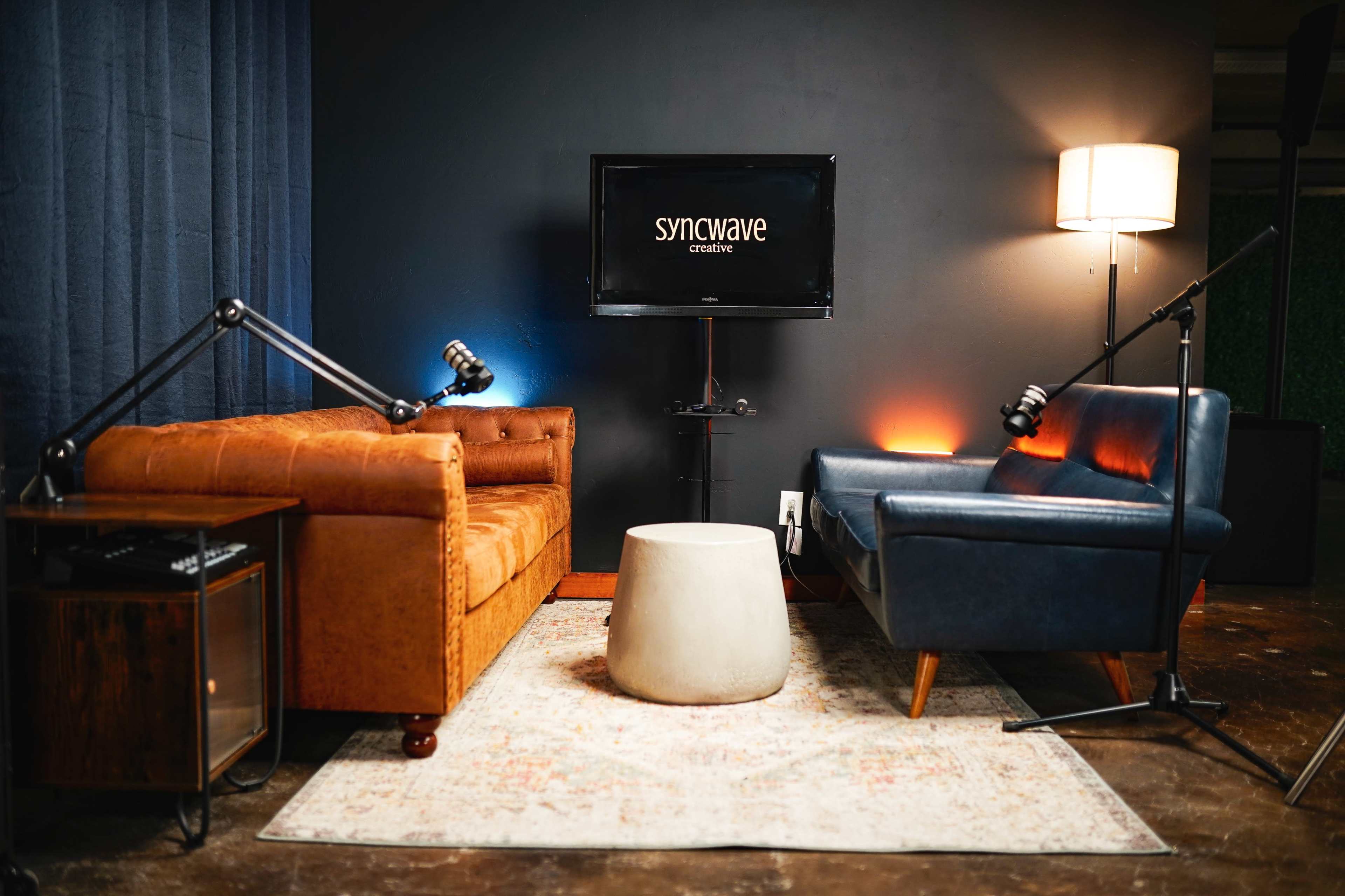 The image shows a modern studio setup featuring two distinct couches, a round concrete table in the center, and a television displaying the "syncwave" logo against a dark wall.