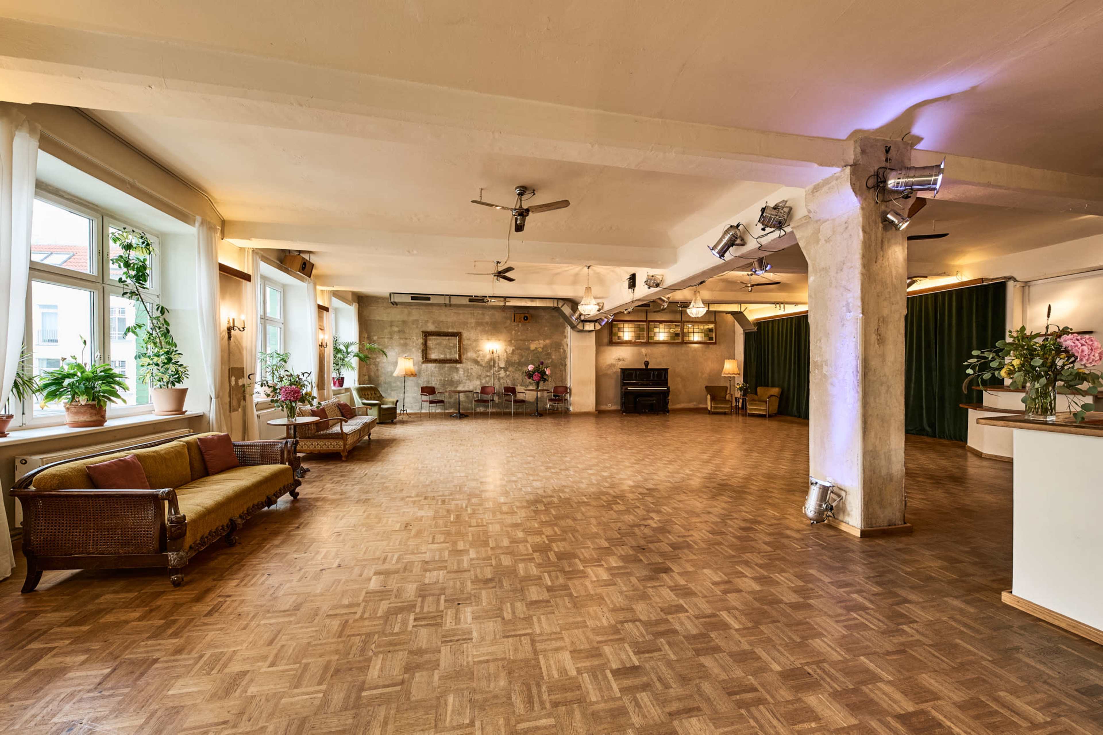 The image shows a spacious, well-lit venue with wooden flooring, large windows, plants, and seating arranged along the walls.