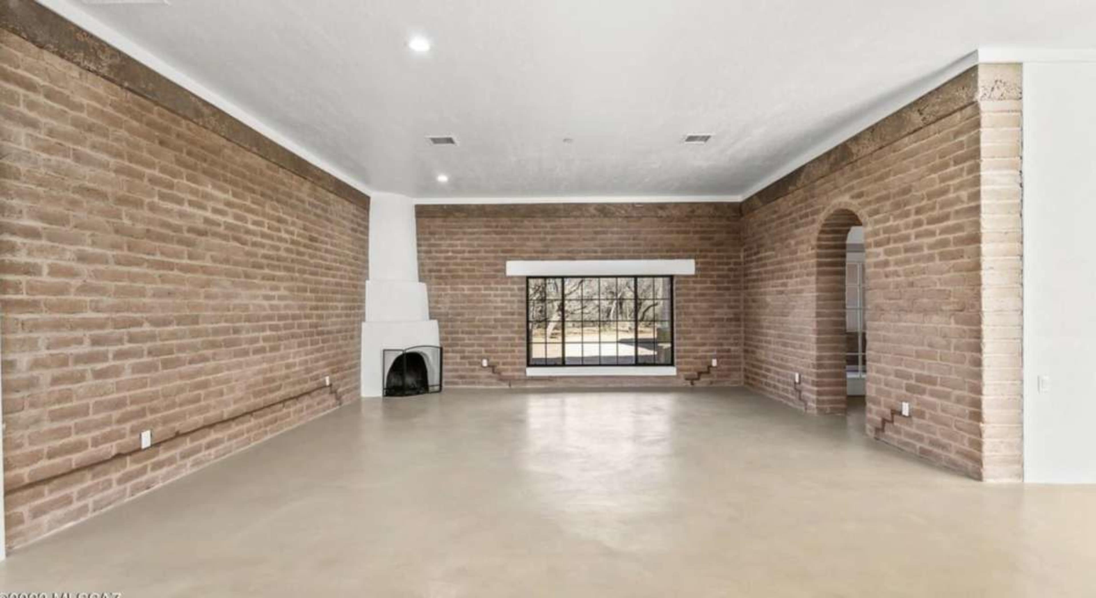 The image shows a spacious interior room with adobe walls, a large window, and a fireplace.